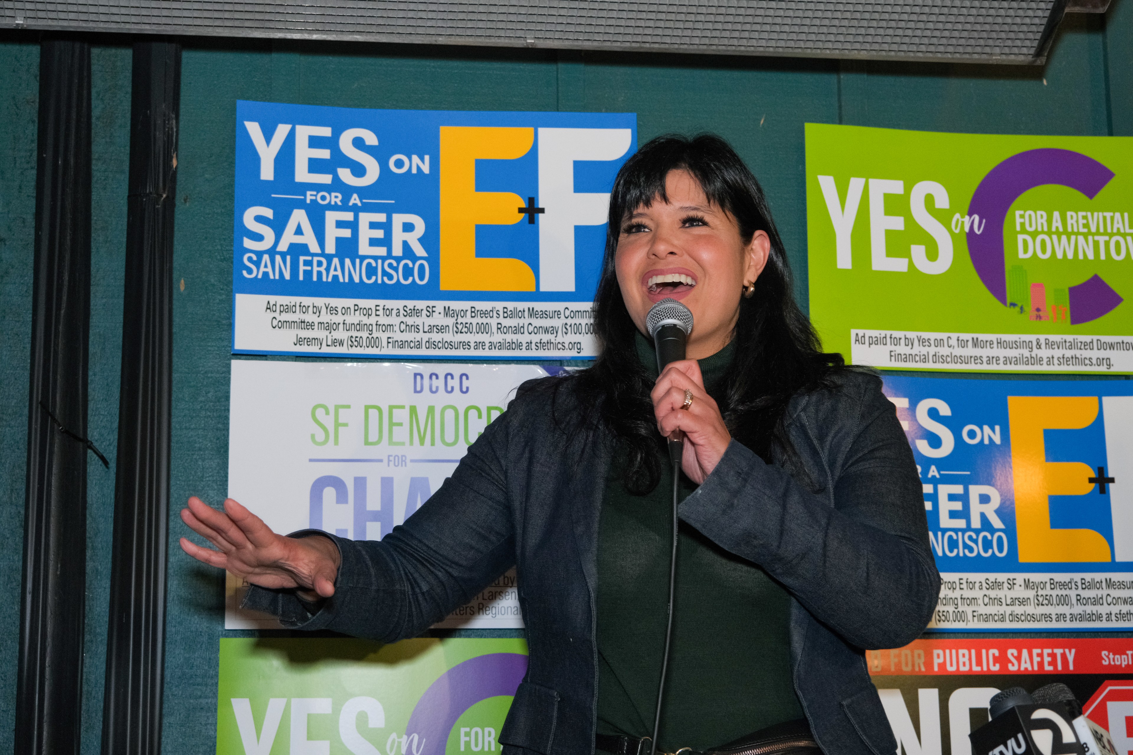 A woman is speaking into a microphone, smiling, with colorful political campaign posters in the background.