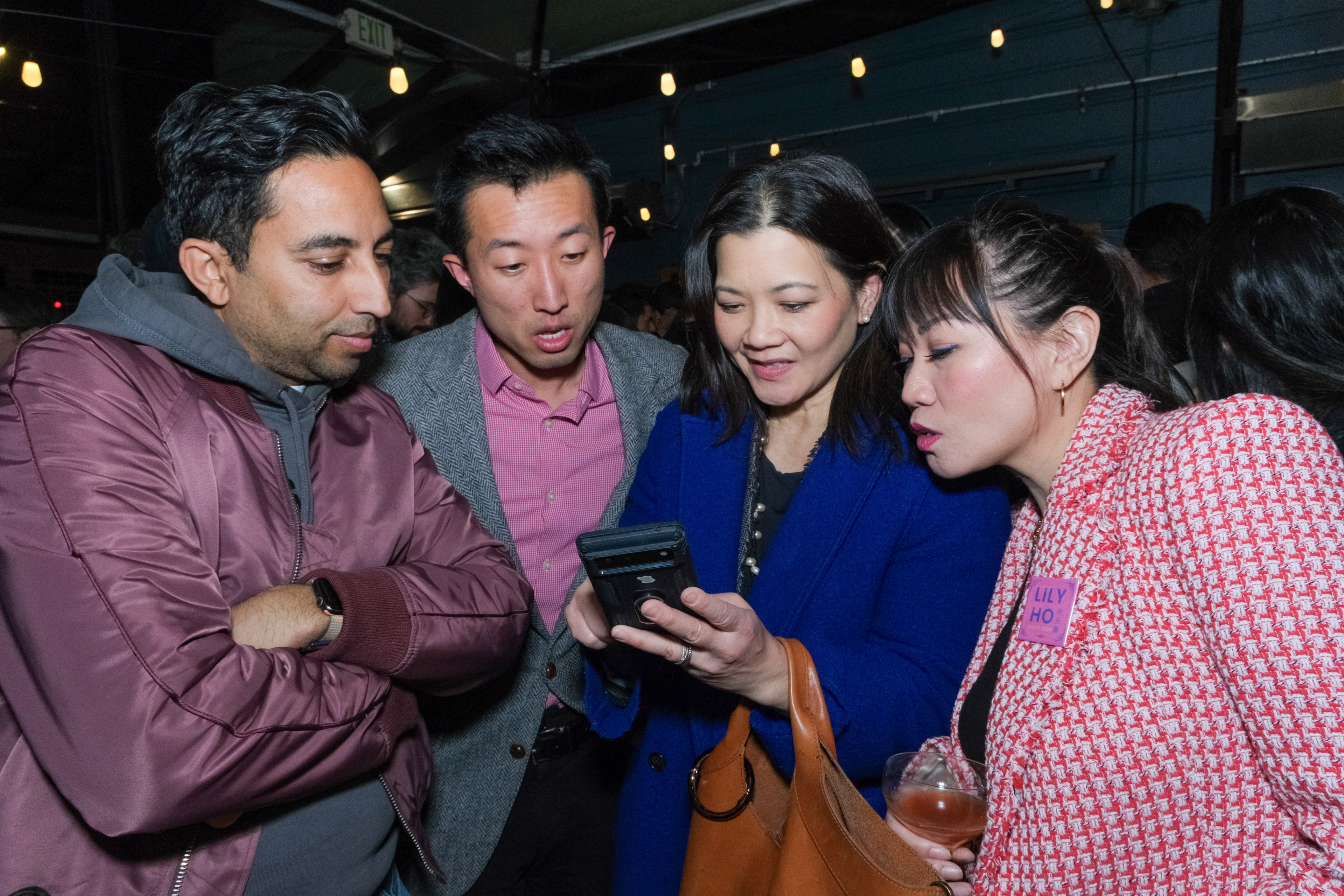 Four people are closely gathered, looking intently at a smartphone that one of them is holding. They appear engaged and curious.