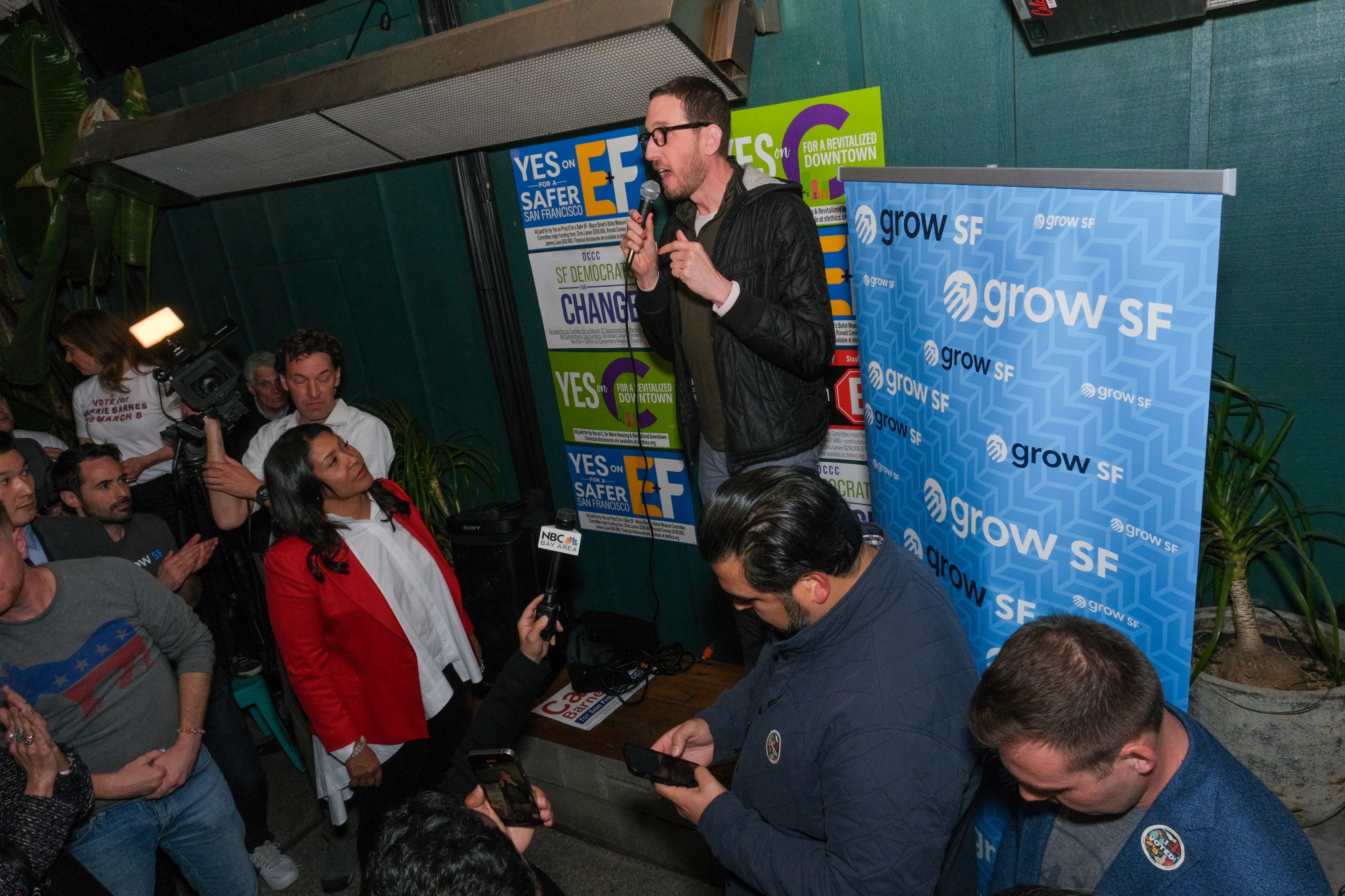 State Senator Scott Wiener speaks into a microphone at a campaign event with onlookers and cameras. Banners read &quot;Grow SF.&quot;