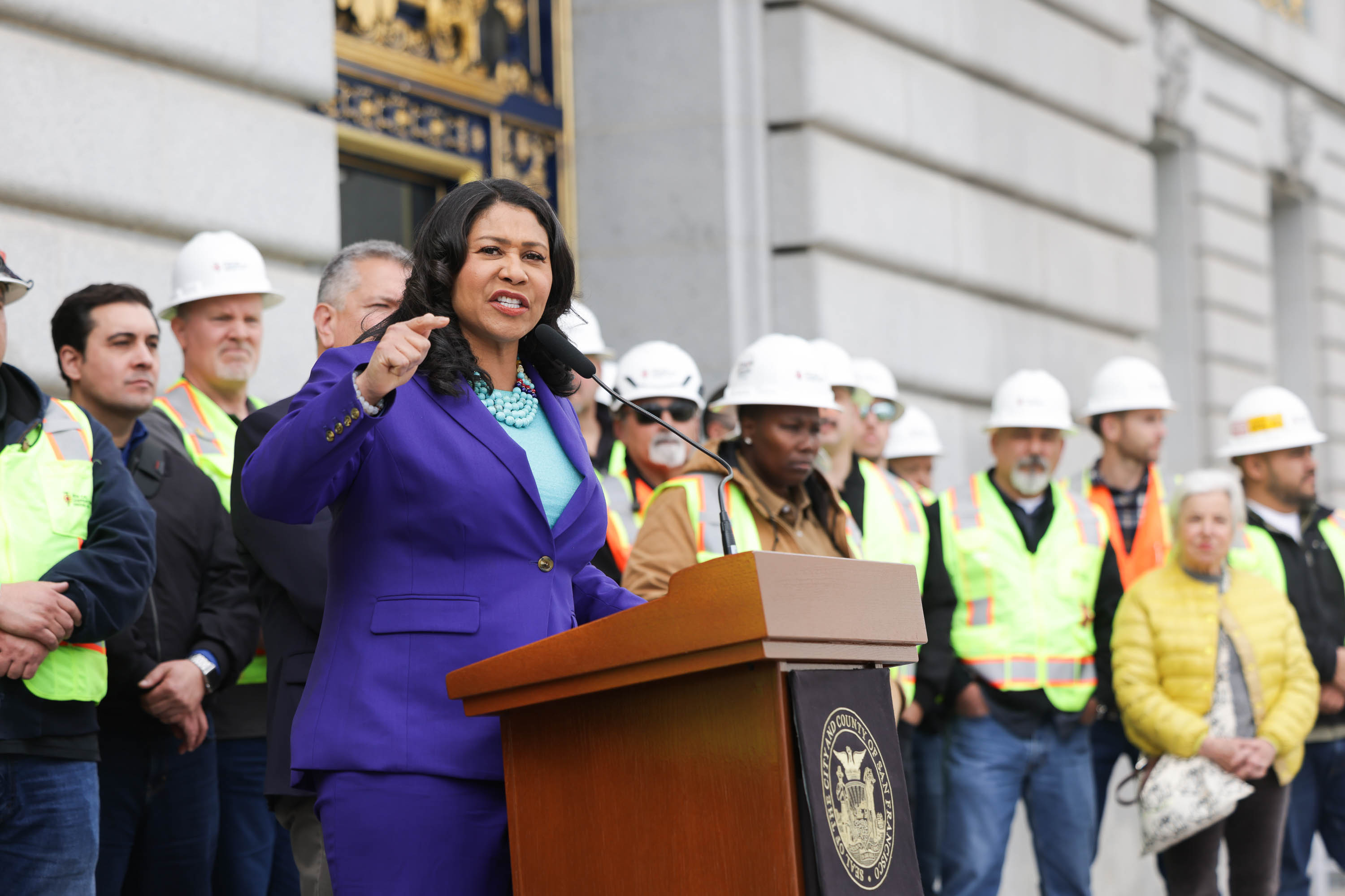 San Francisco Mayor London Breed speaks at a podium with a group of people in construction vests and helmets.