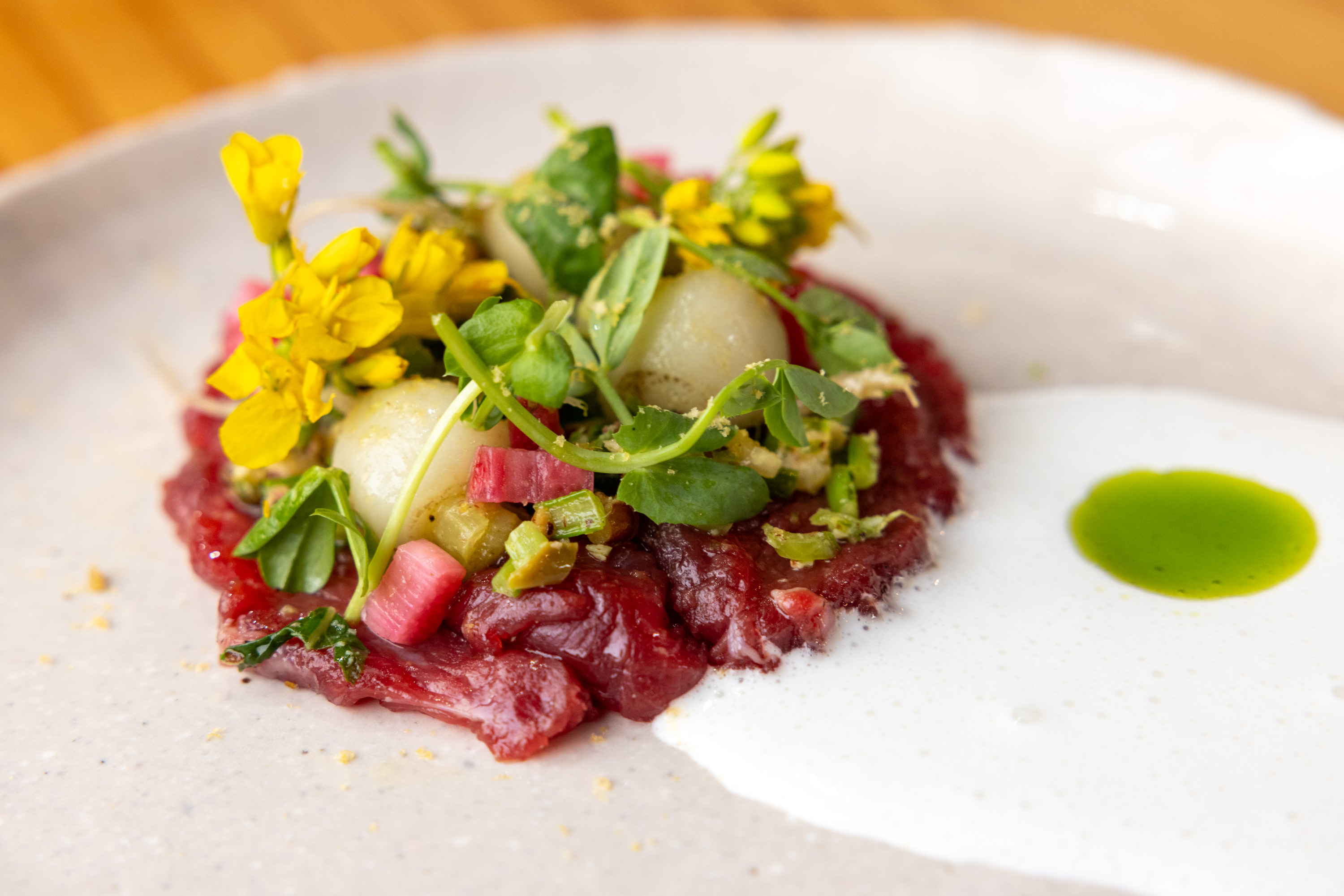 A plate of beef tartare topped with green apple, herbs, edible flowers, and rhubarb, with a drizzle of green sauce.