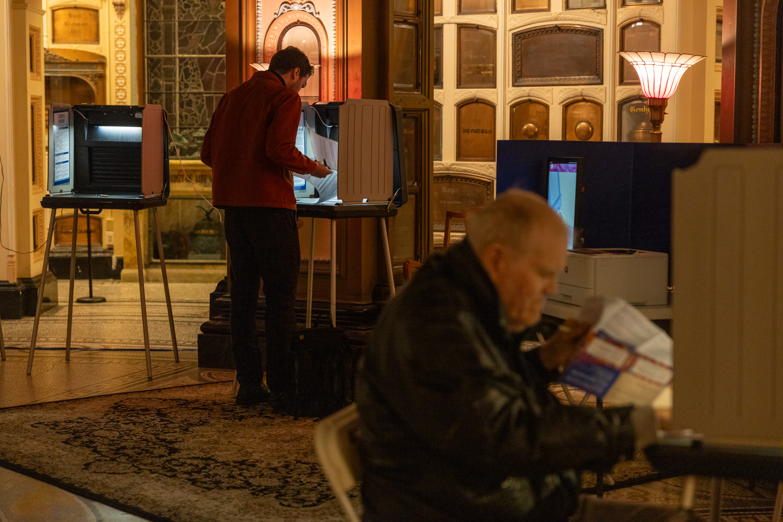 A man in red votes at a booth while another reads a paper in an ornate room with vintage decor.