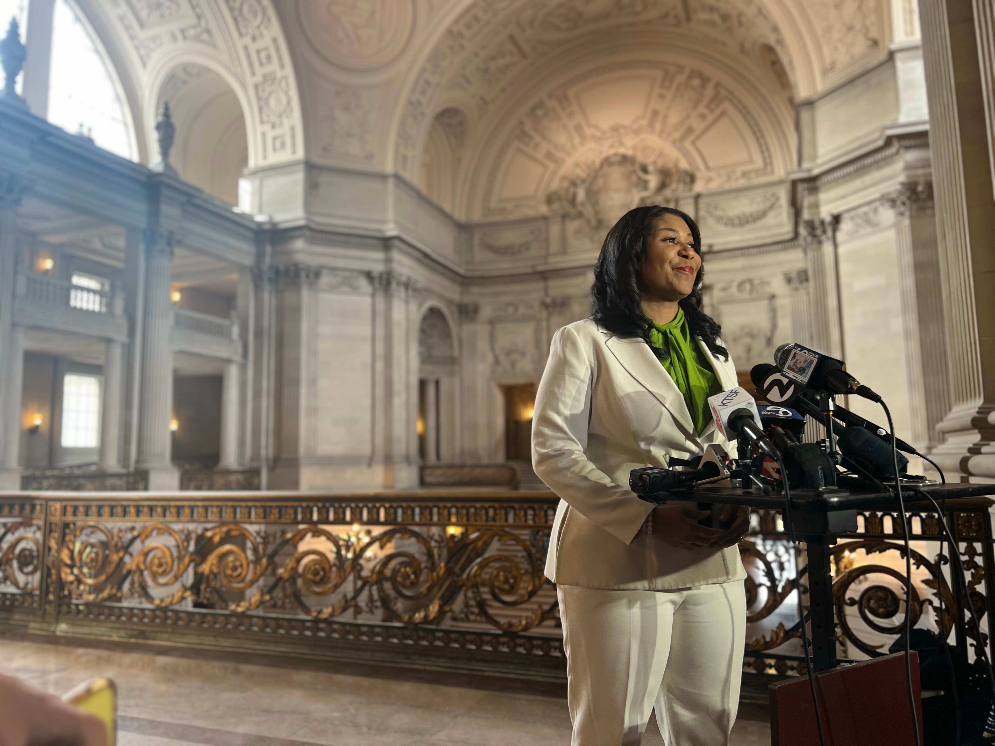 A woman in a white suit speaks at a podium with microphones, inside a grand hall with ornate architecture.