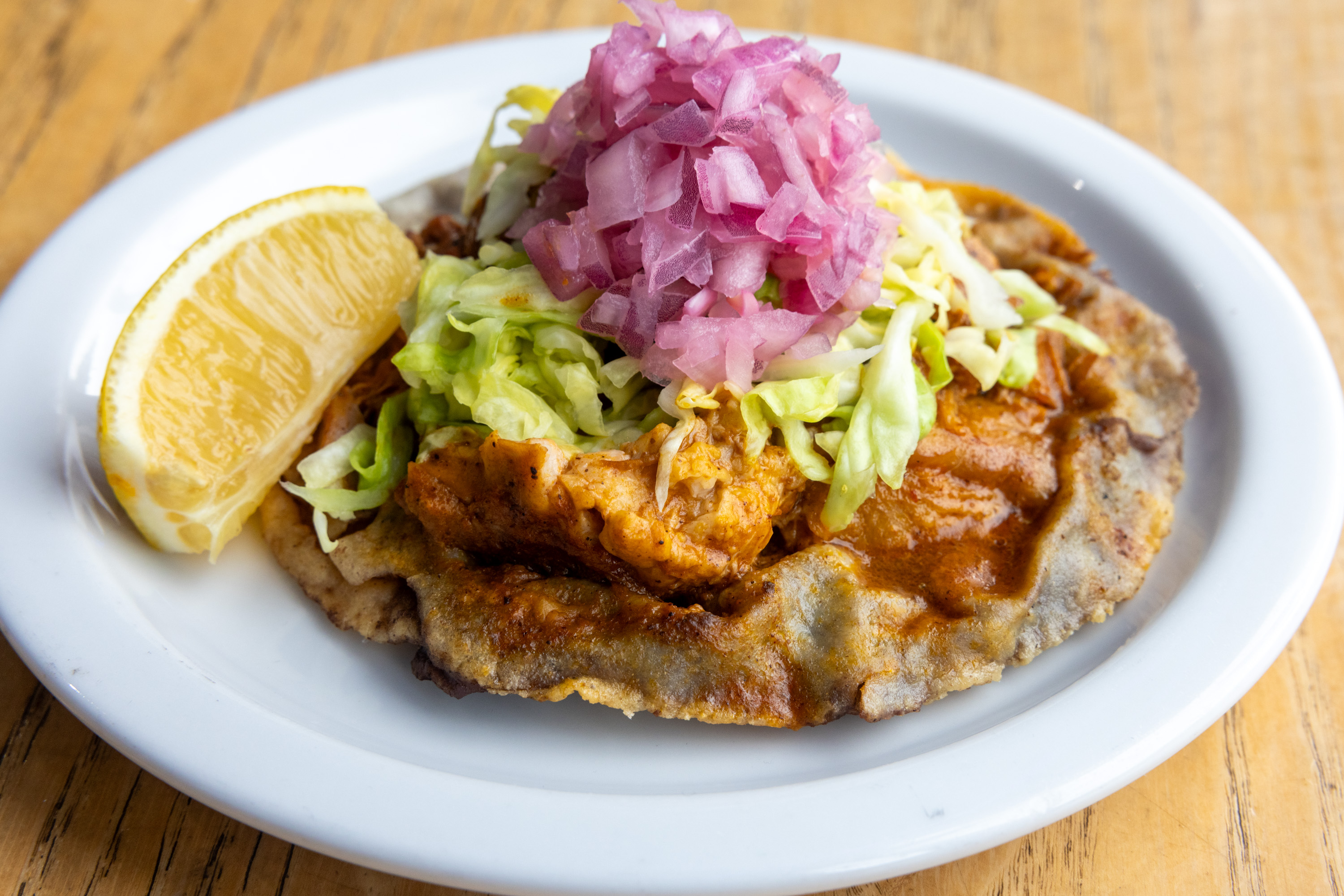 A taco with chicken, lettuce, pickled onions, and a lemon wedge on a white plate.