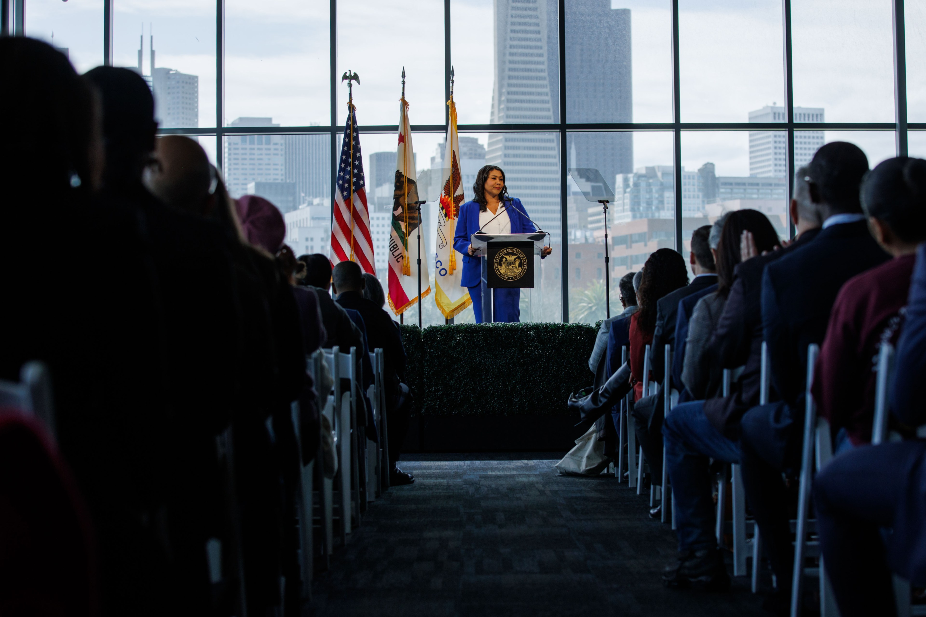A person at a podium speaking to an audience with city skyline visible through floor-to-ceiling windows in the background.