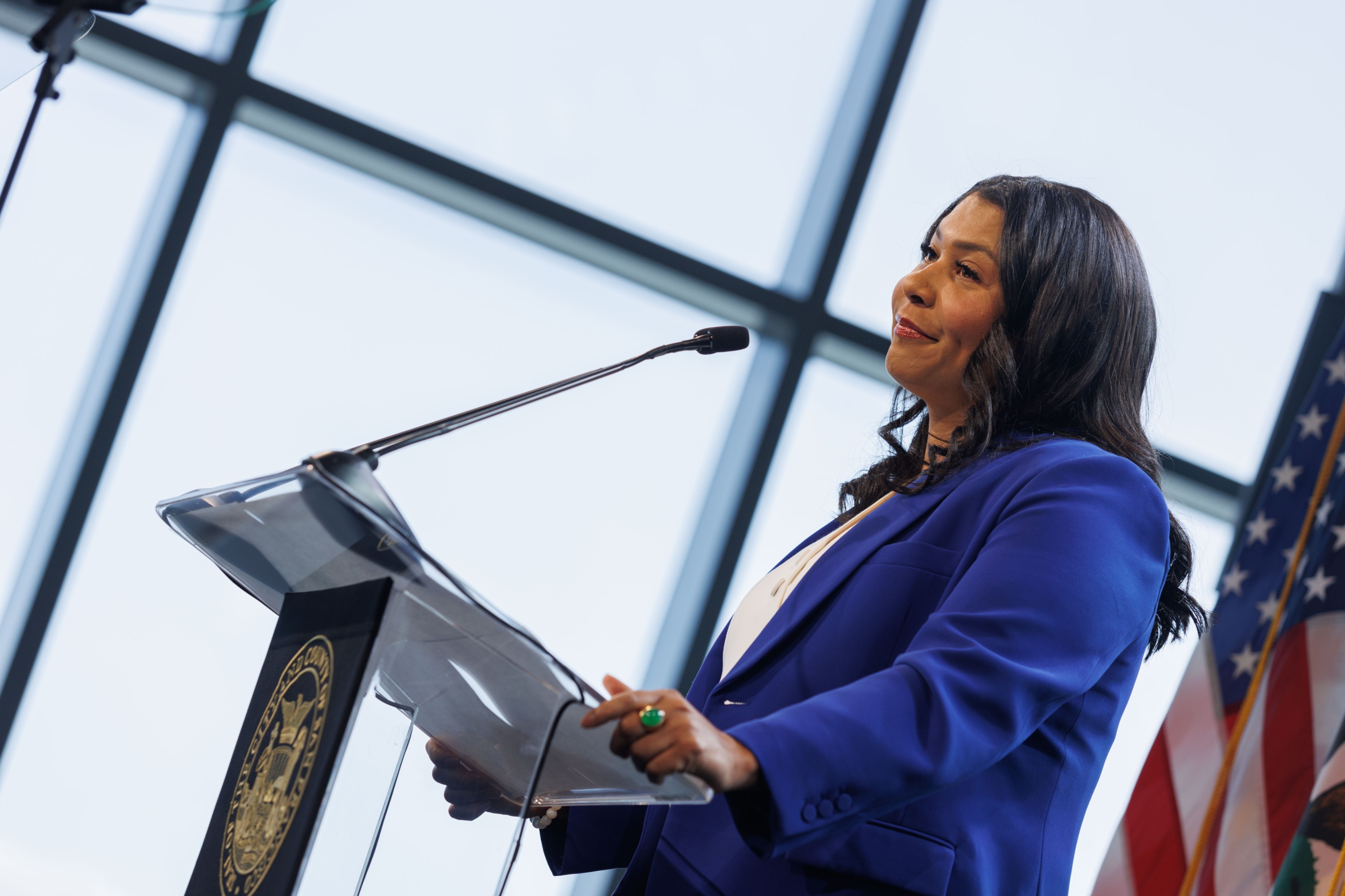 Mayor London Breed in a blue blazer speaks at a podium with a mic, behind her is a window and U.S. flags.