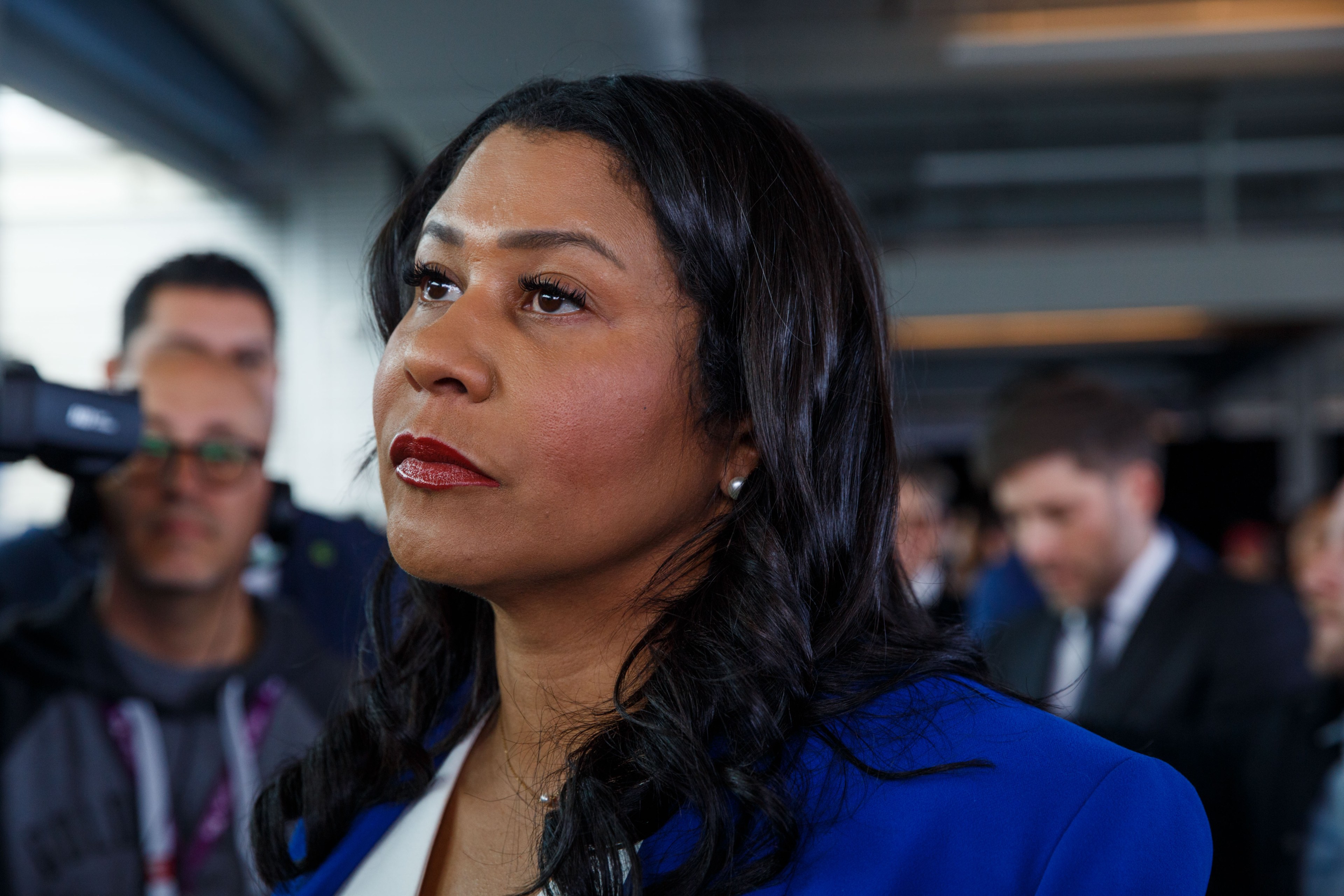 Mayor London Breed in a blue blazer appears focused, with people and a camera in the blurred background.