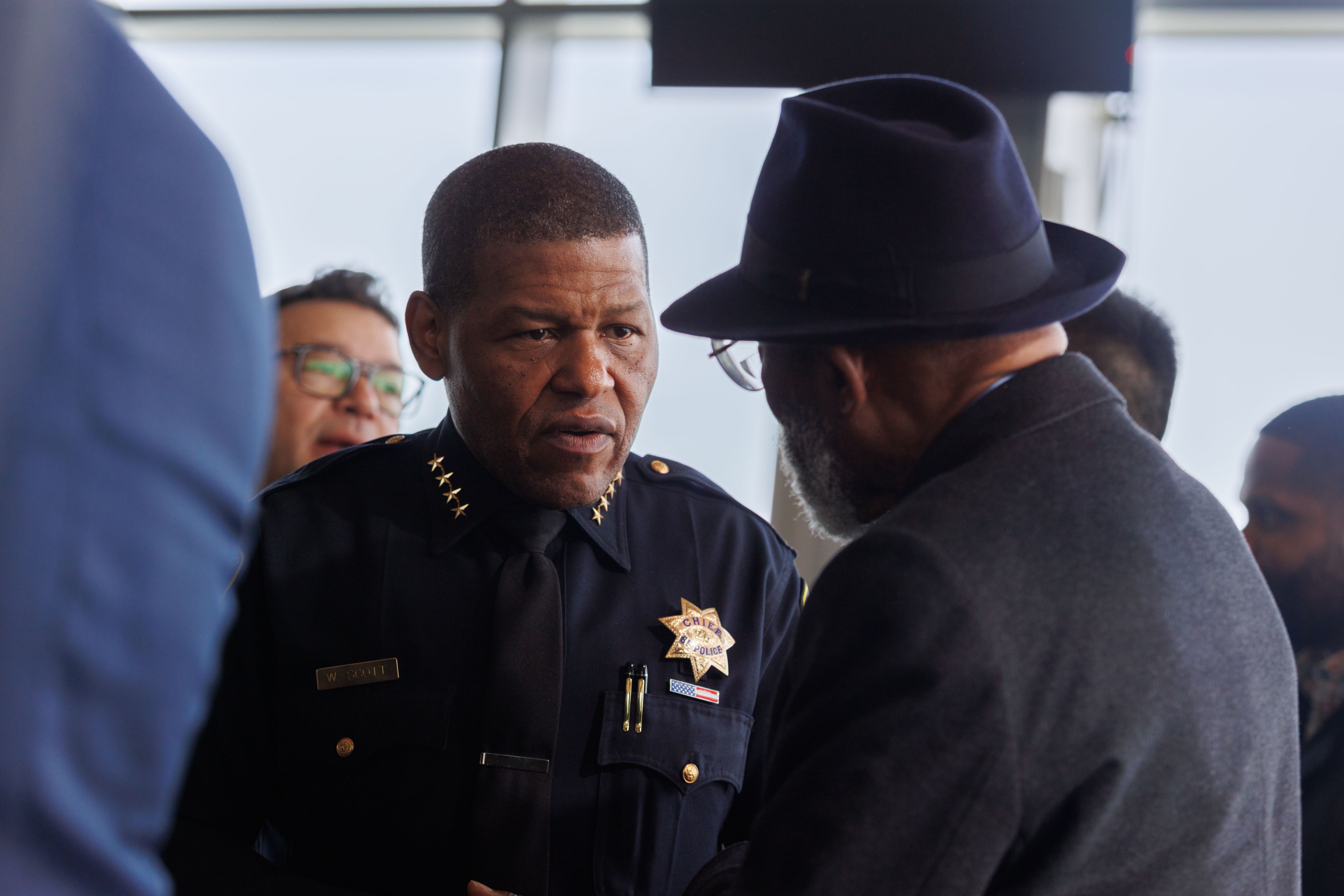 A police chief in uniform converses with a man in a black hat and coat.