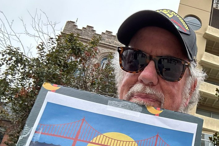 Richard Parina, who has a beard and wears sunglasses and a cap, holds up a picture of the Golden Gate bridge with trees.