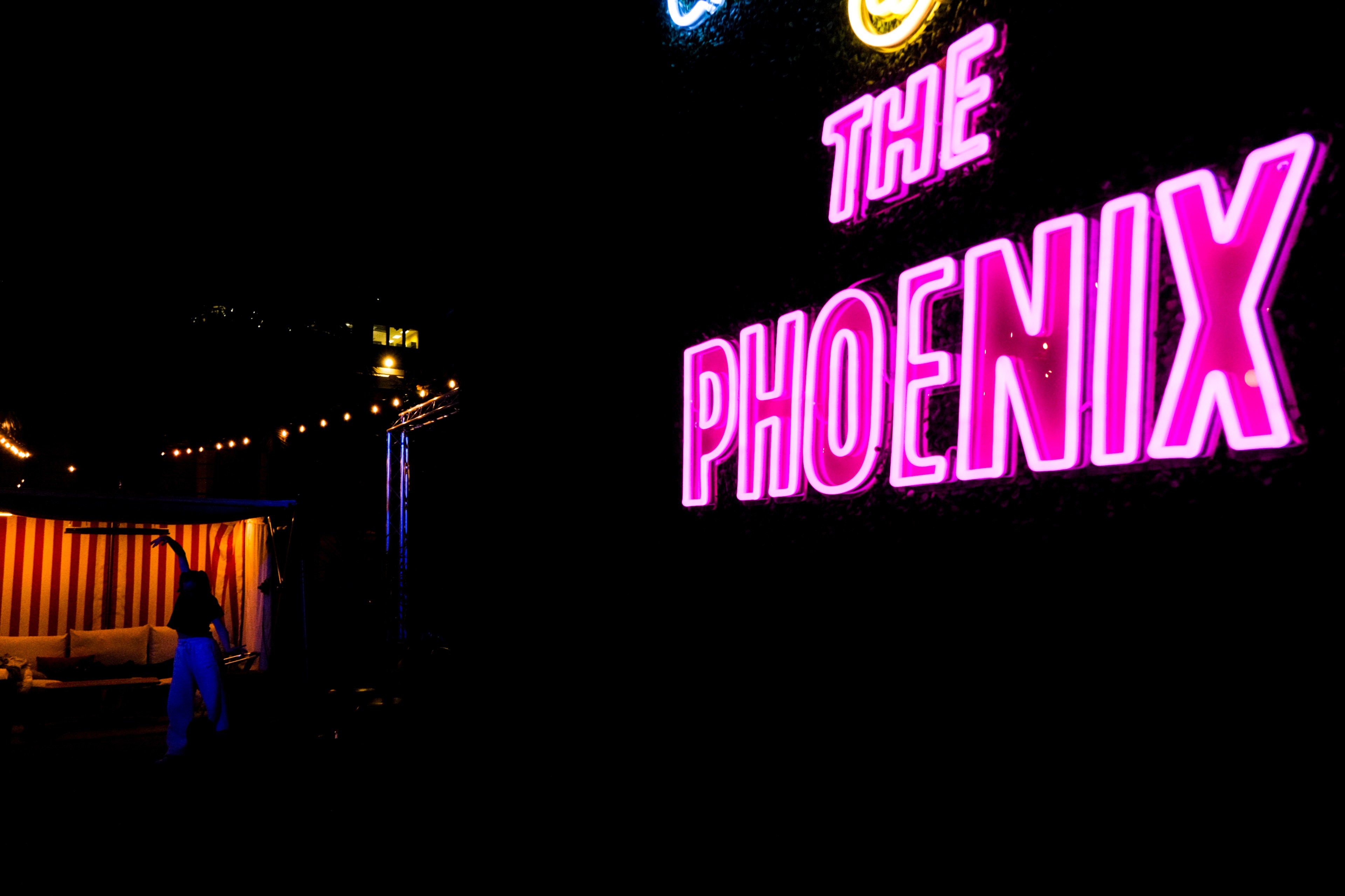 Neon sign reading &quot;THE PHOENIX,&quot; a lit-up tent with a person, and string lights against a dark backdrop.