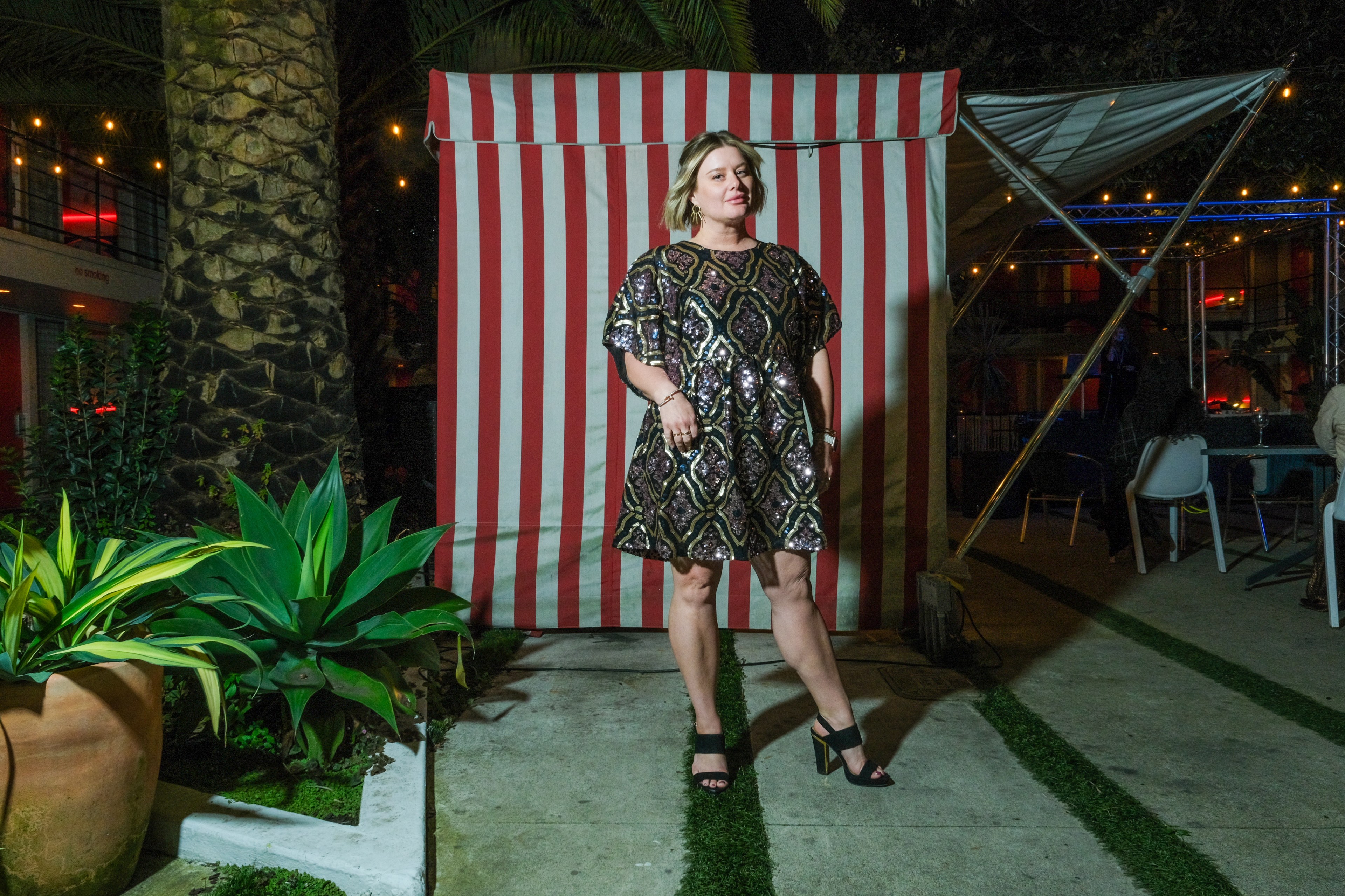 A woman poses before a striped backdrop at a nighttime outdoor event, with plants and ambient lighting around.