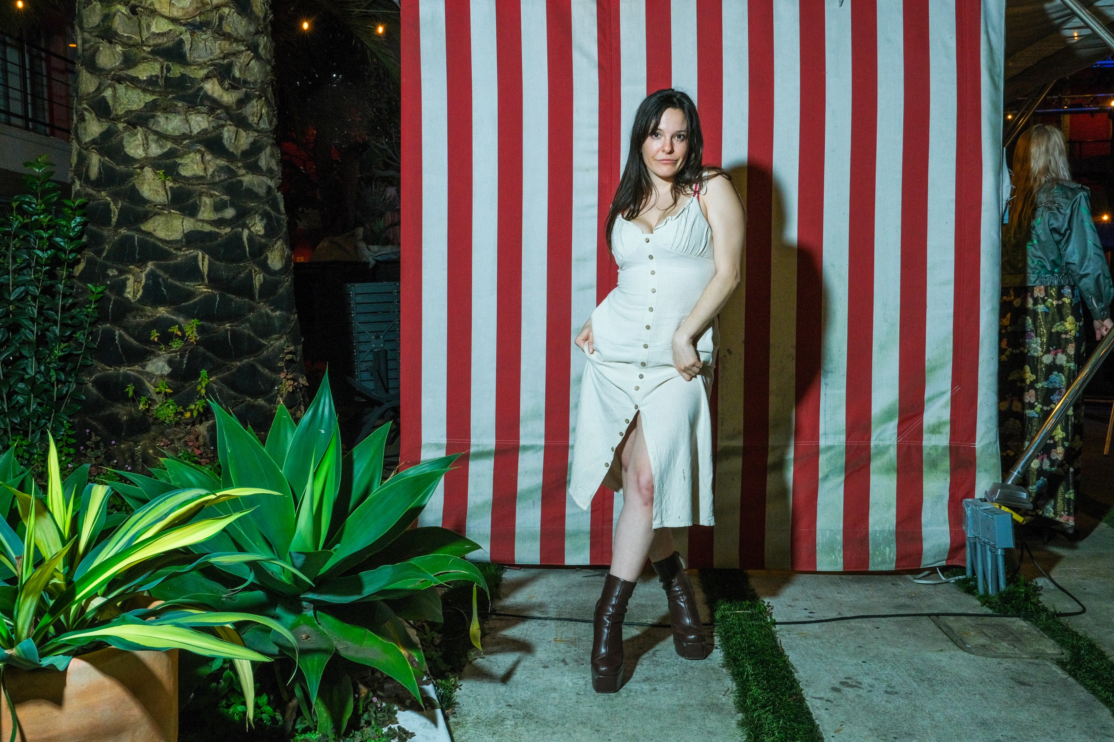 A woman poses in front of red and white stripes, wearing a white dress and brown boots, with plants nearby.