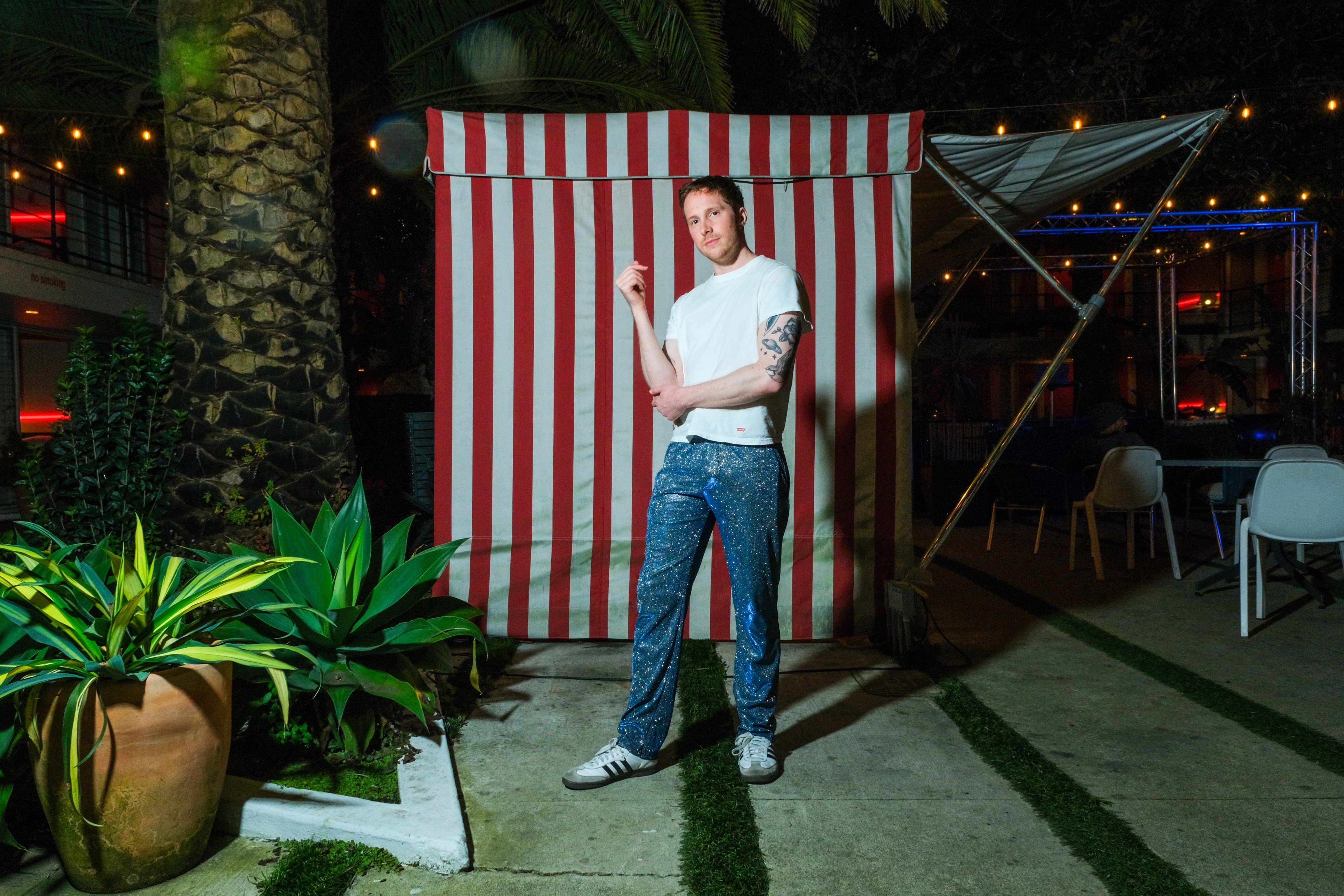 A man poses before a red-striped backdrop at a night event, sporting a white tee and patterned pants.