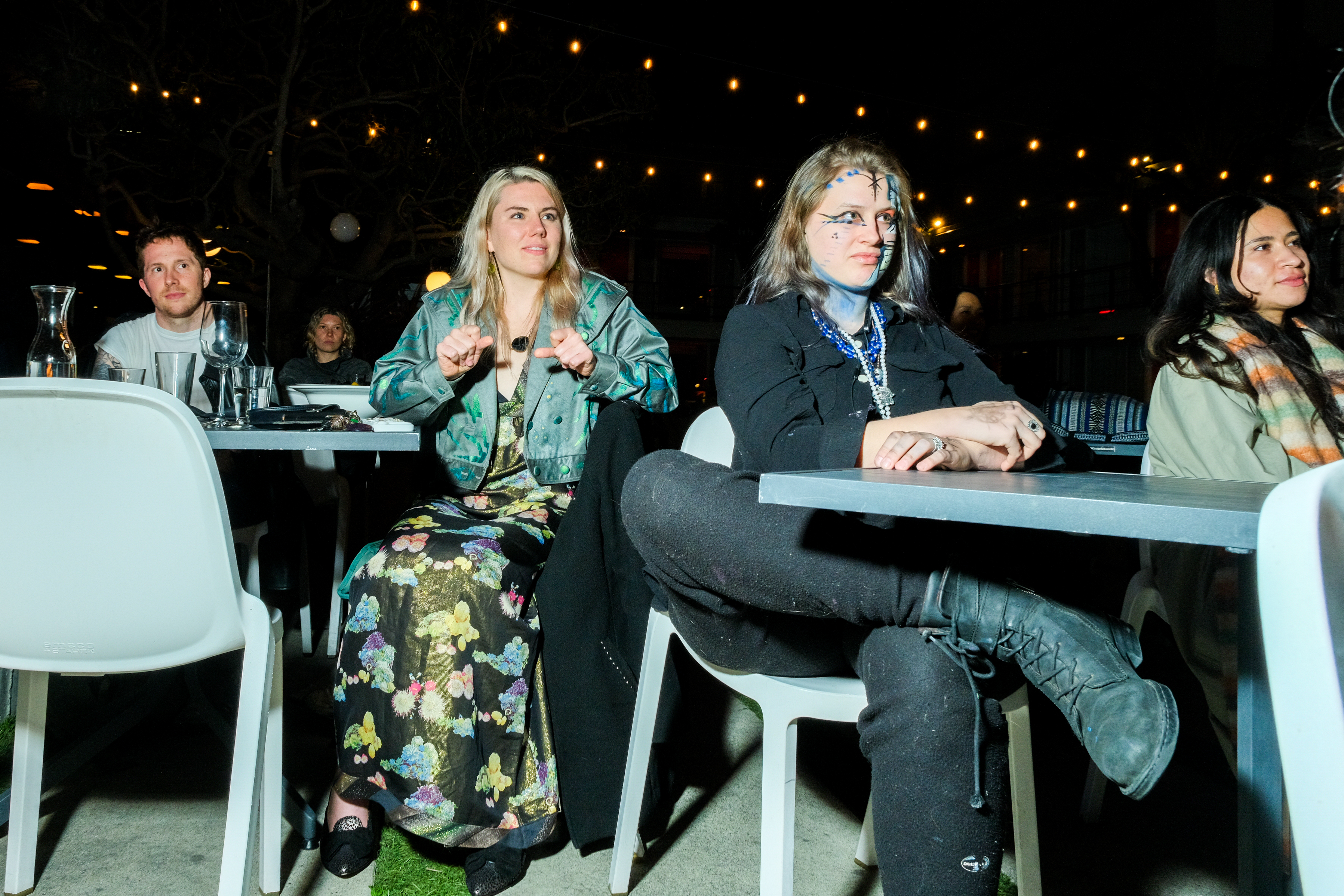 Four people sit at an outdoor table at night, with string lights above and half-empty glasses.