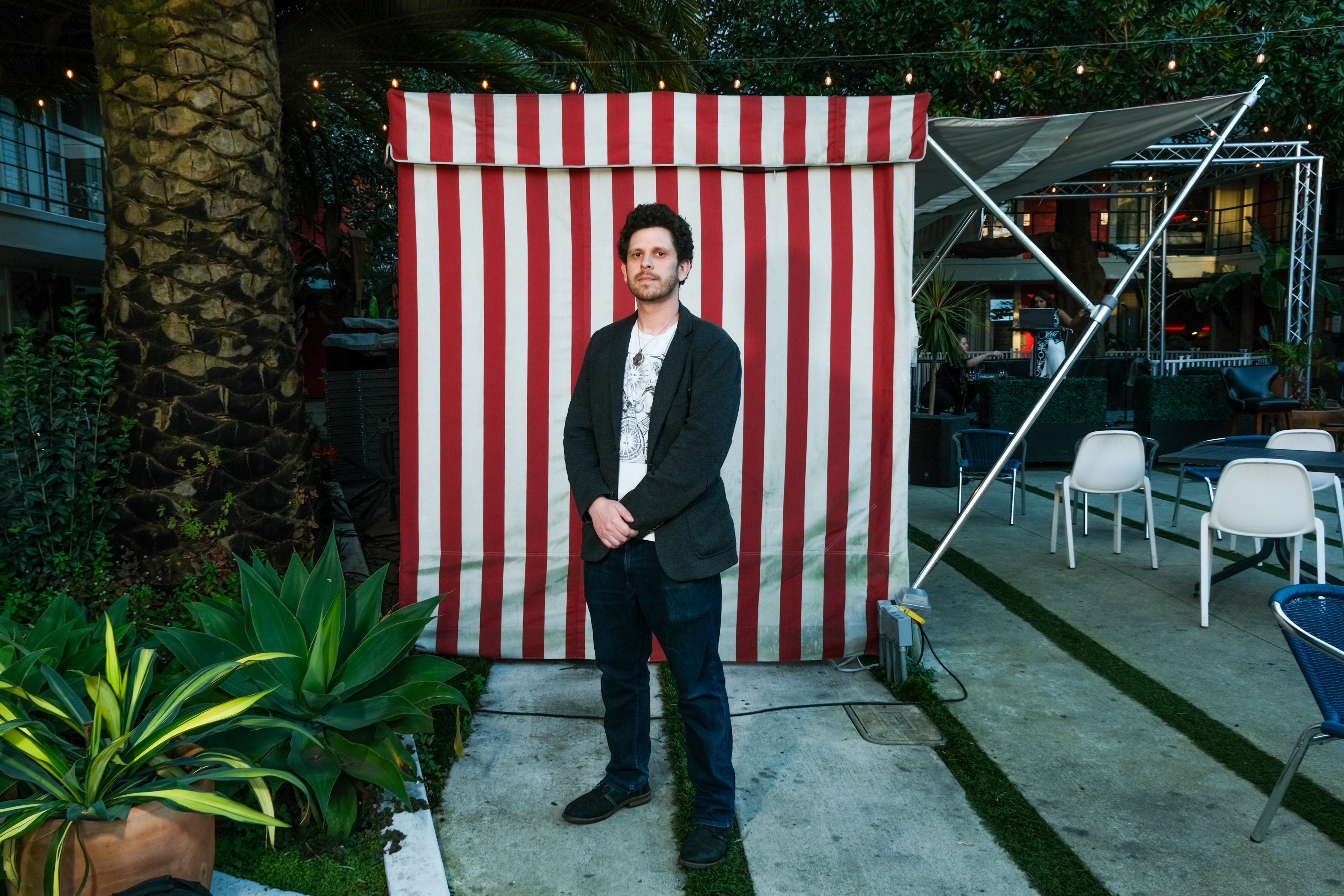 A man stands in front of a red and white striped backdrop outside with plants and chairs around.