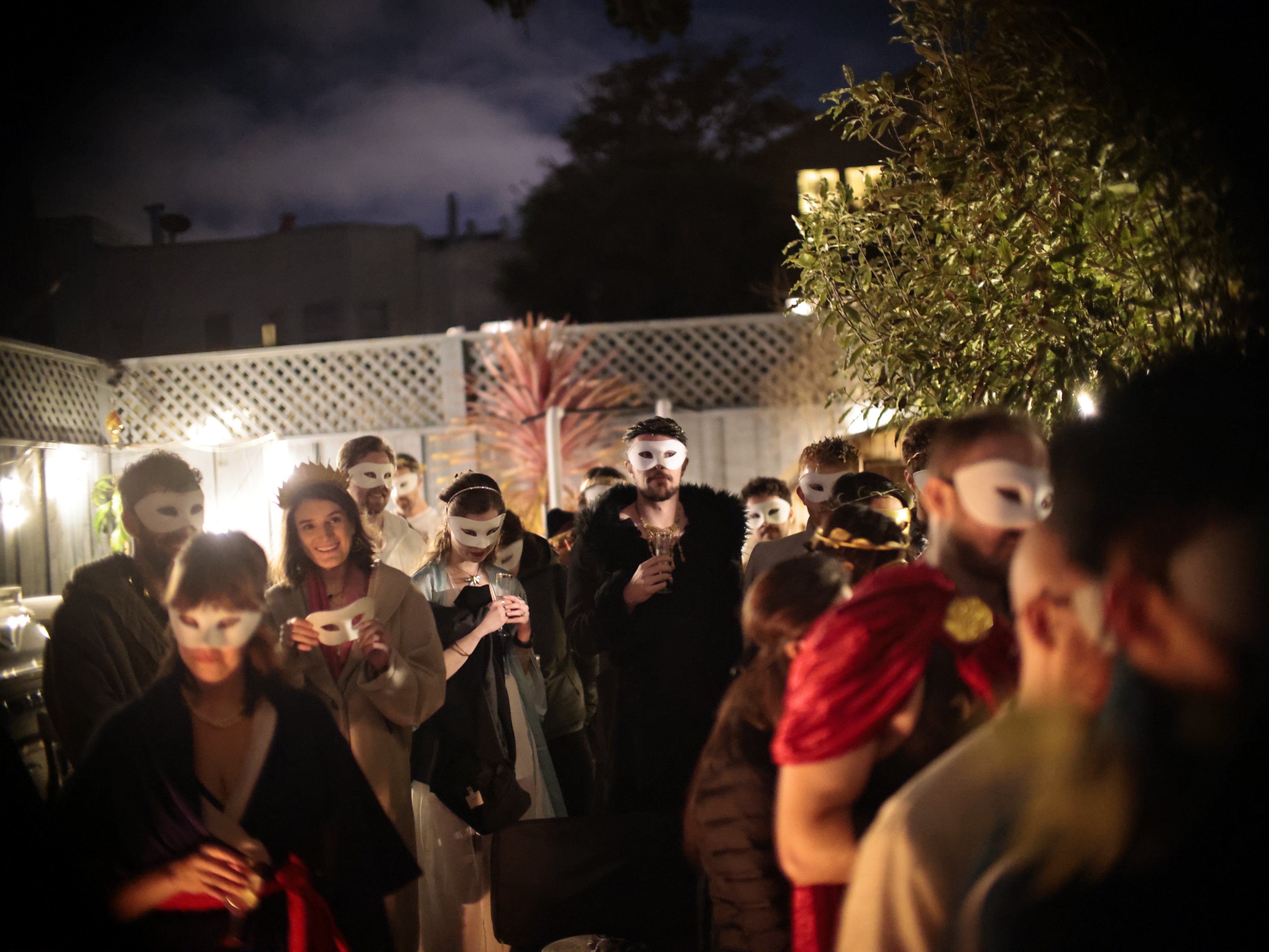 A nighttime gathering of people wearing masks, with soft lighting and a tree in the background.