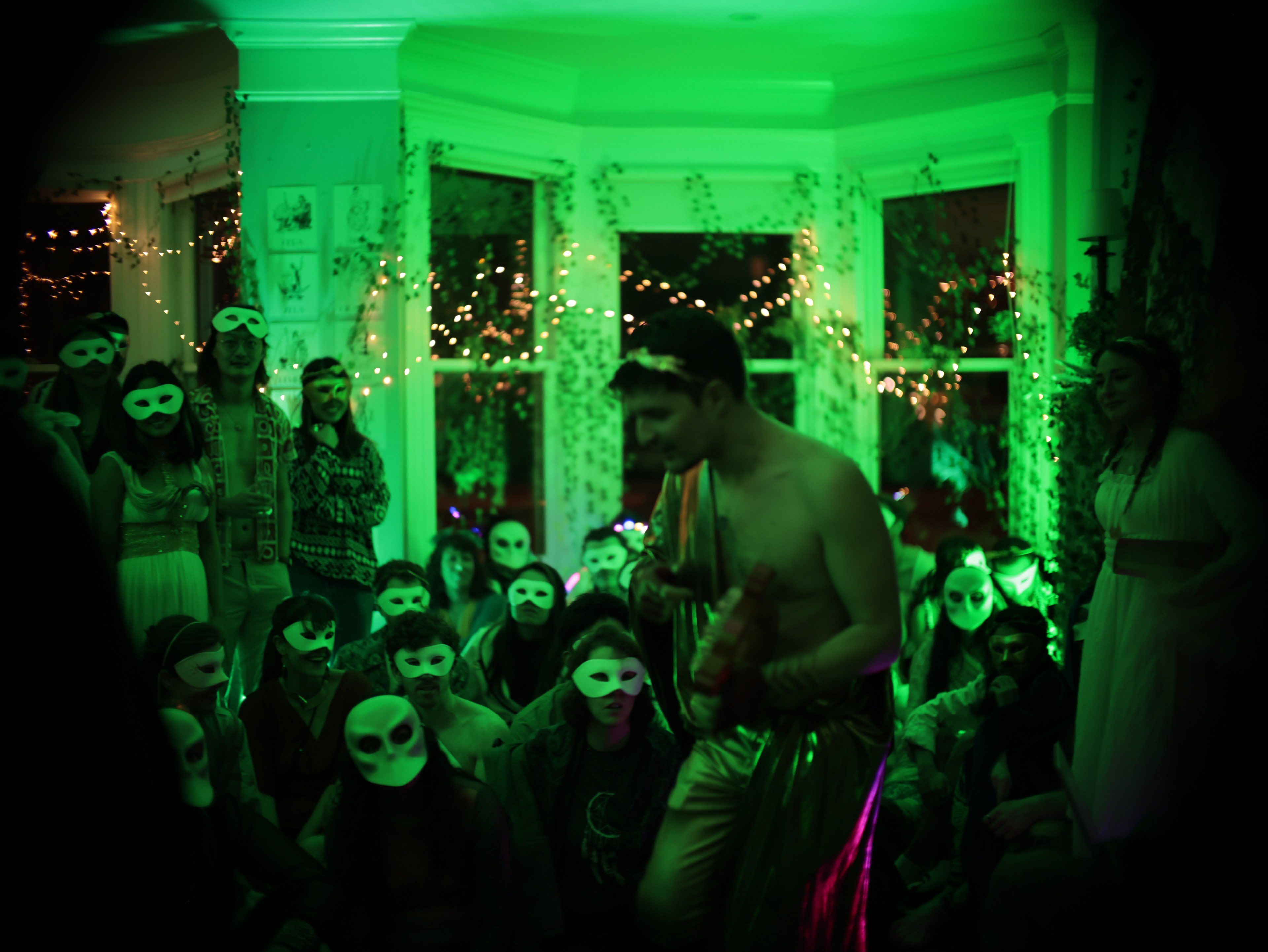 A dimly lit room with masked people watching a shirtless man in golden pants under greenish lighting with string lights.