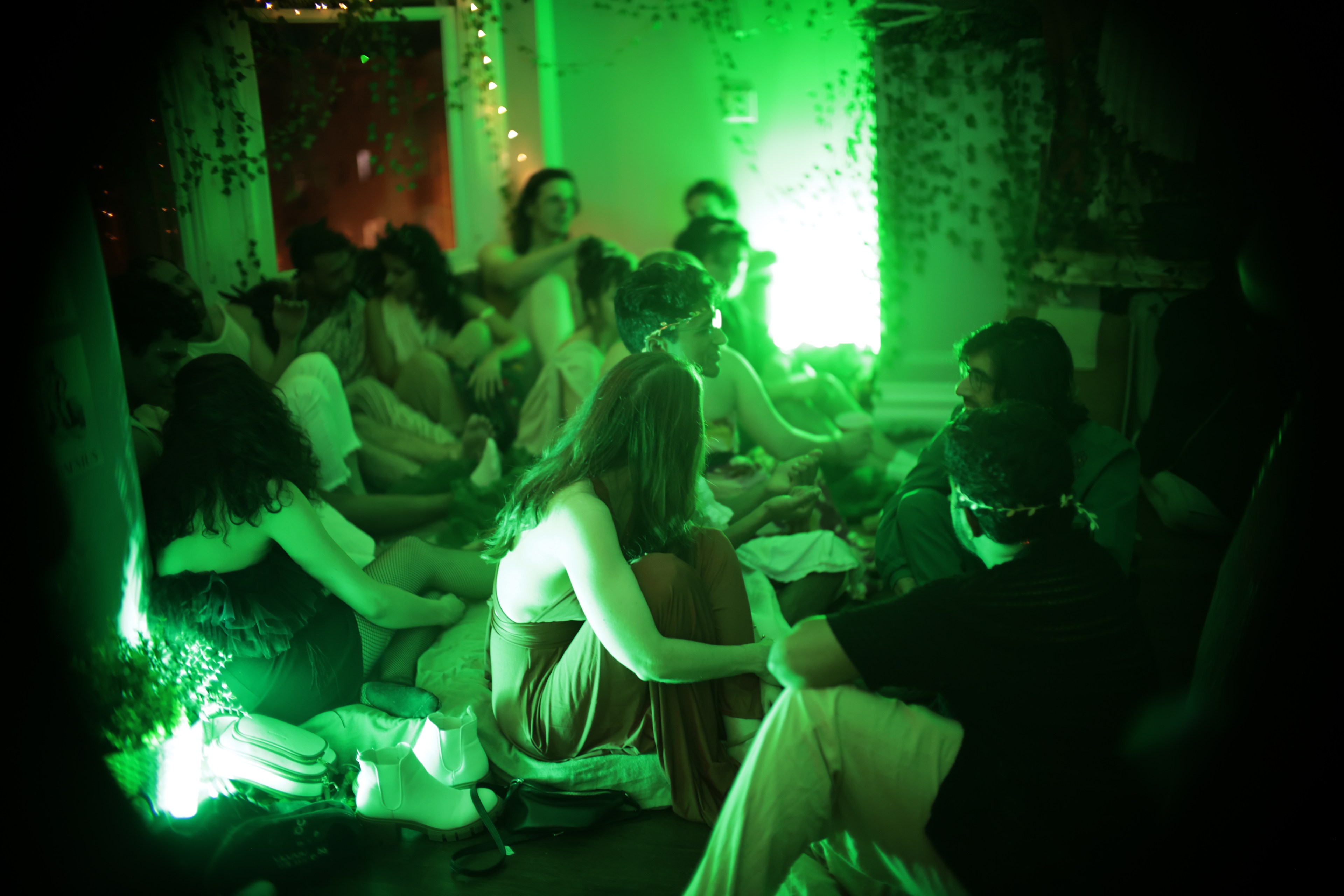 A group of people sit on the floor in a room with green lighting and fairy lights, chatting and relaxing.