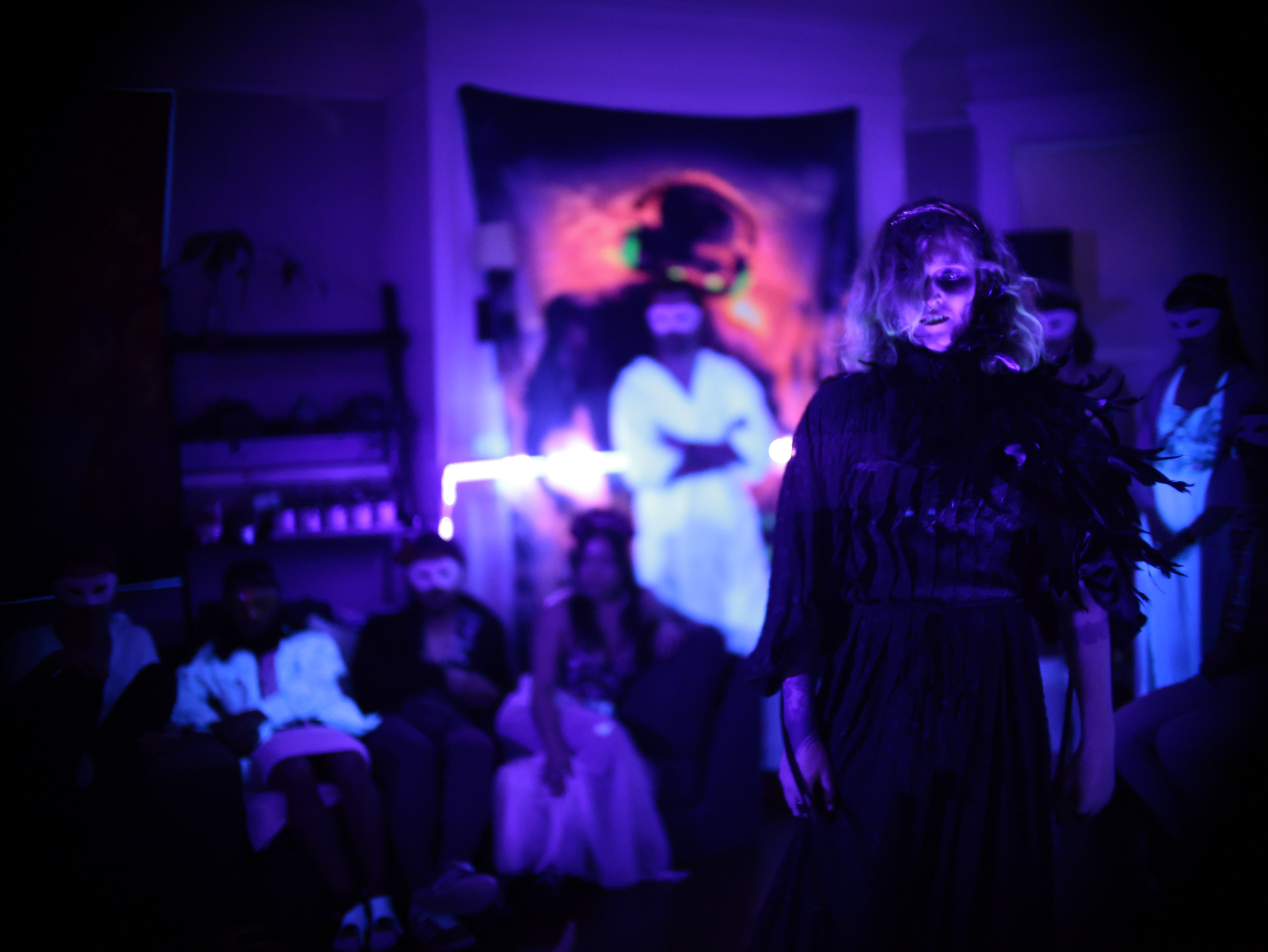 One person in spooky attire stands in a dimly lit room with people in costume. Purple lighting sets a haunting mood.