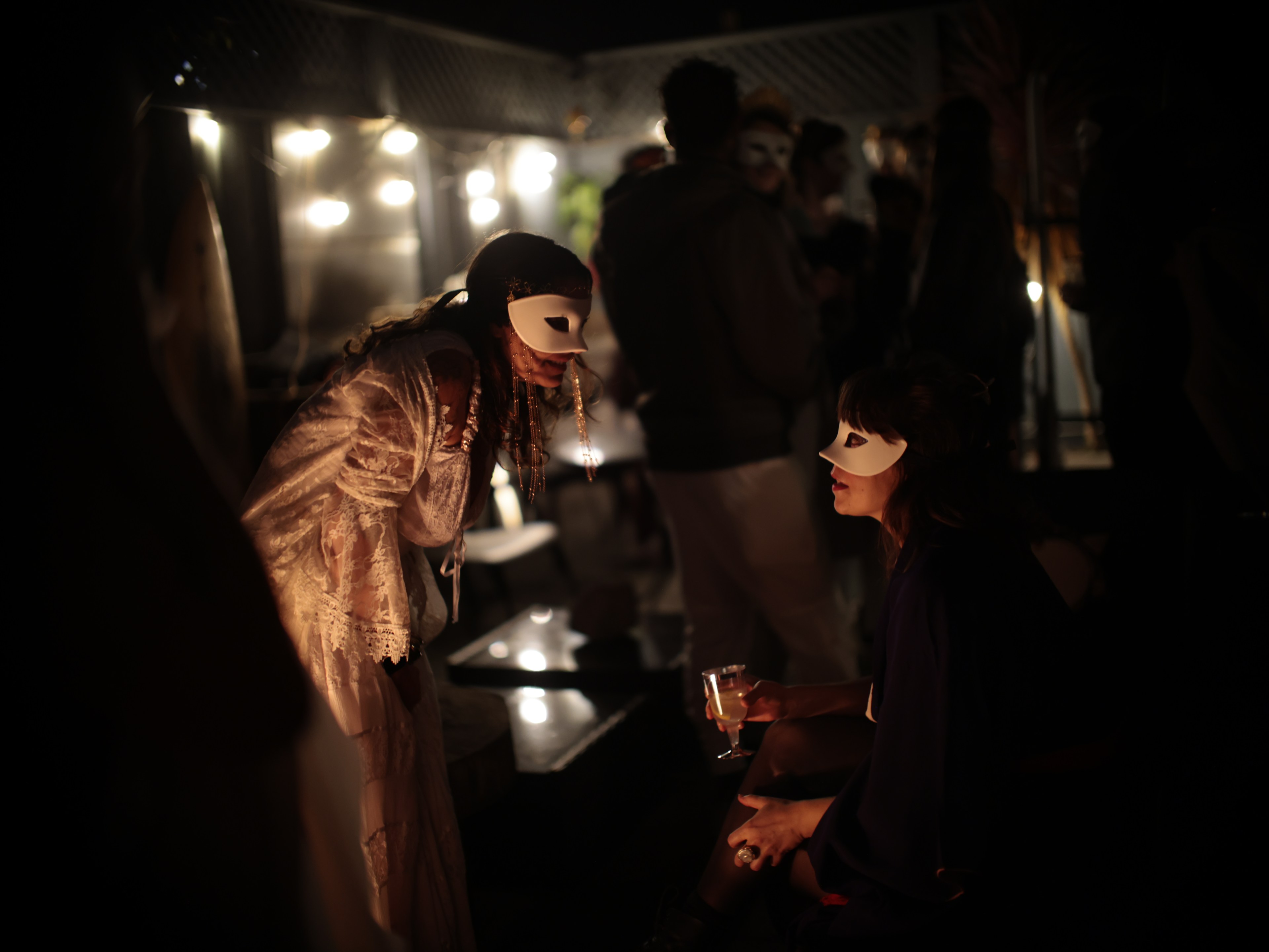 Two people wearing masks talk at a dimly lit party; one is seated with a drink, the other stands leaning in.