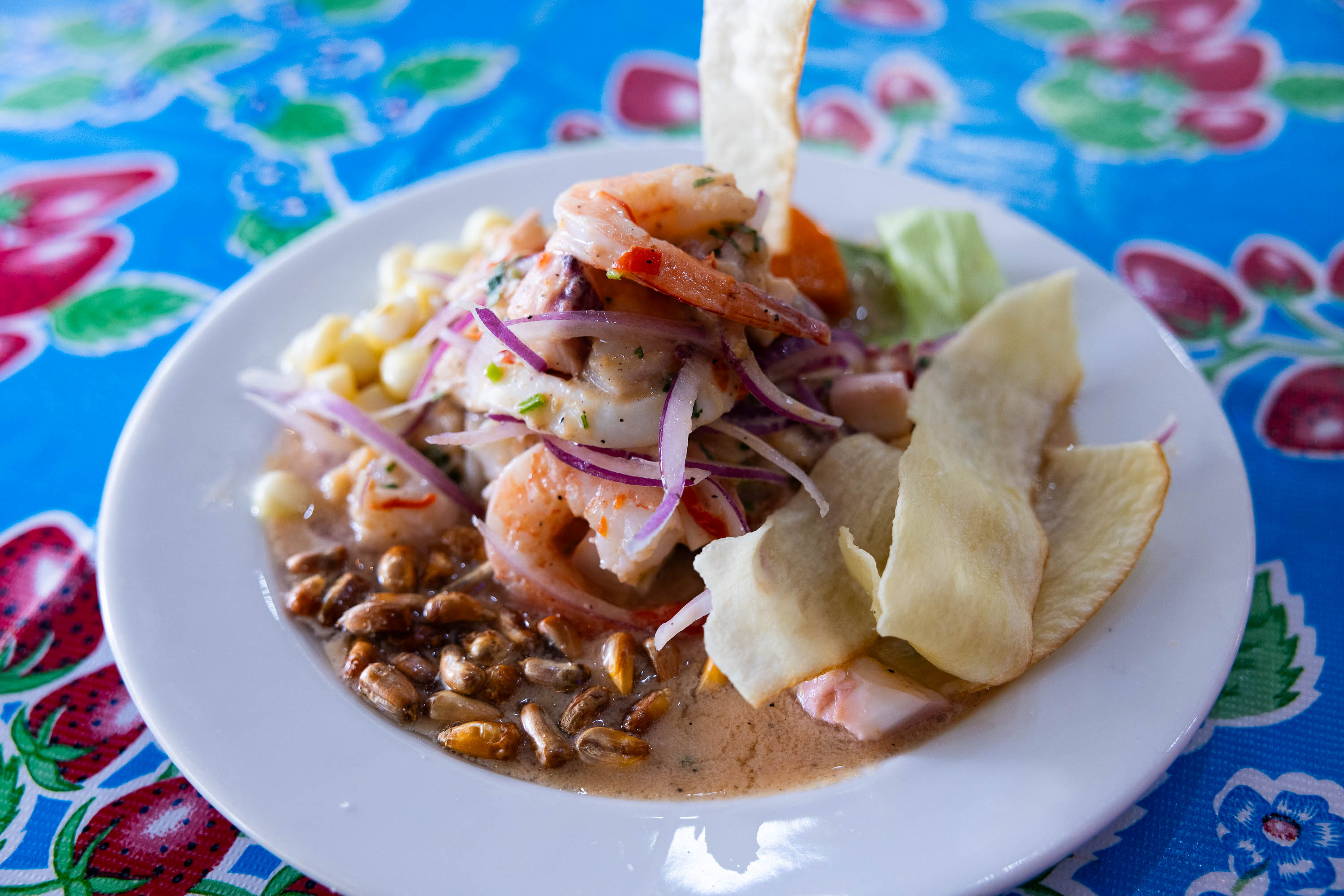 A plate of ceviche with shrimp, corn, onion, and fried plantains on a blue floral tablecloth.