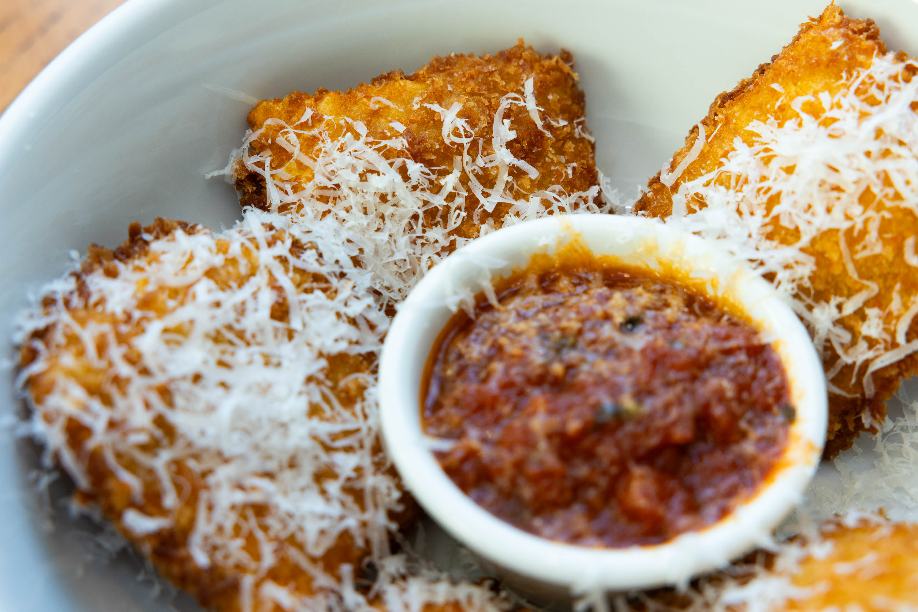 Fried ravioli topped with grated cheese served with marinara sauce in a white bowl.