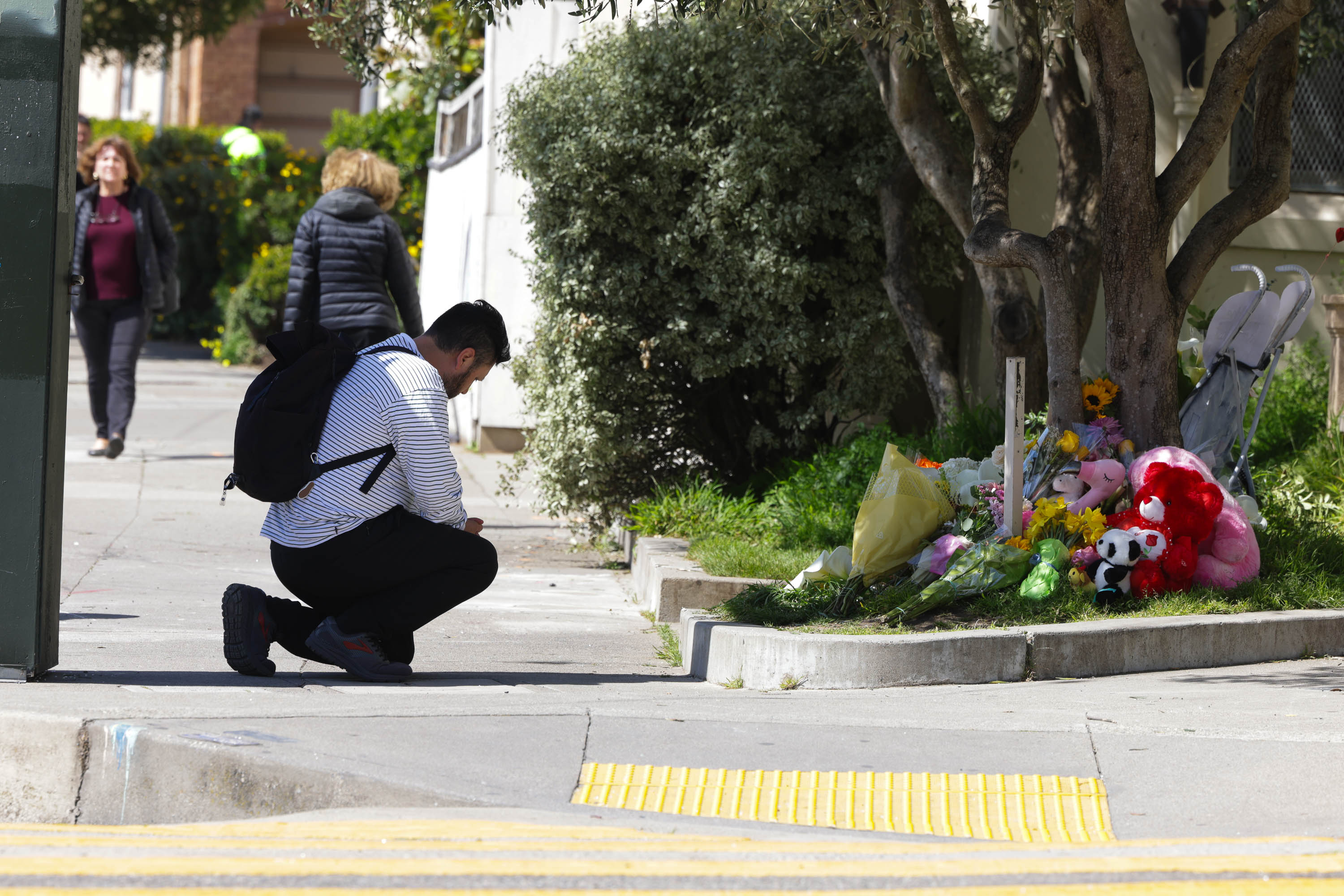A person crouches by a sidewalk memorial with flowers and stuffed animals, as another person walks by in the background.