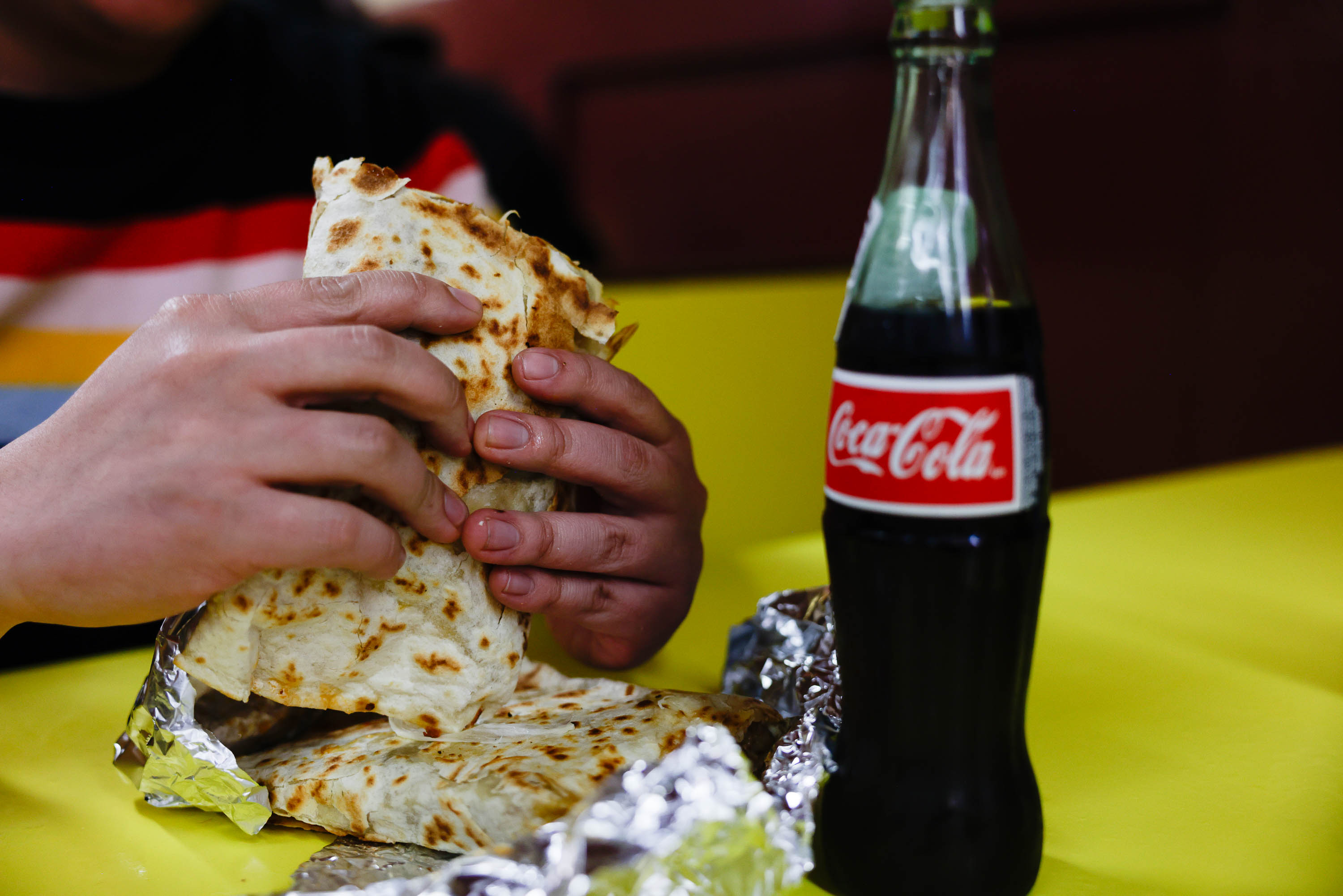 Hands holding a quesadilla with a Coca-Cola bottle on a yellow table.