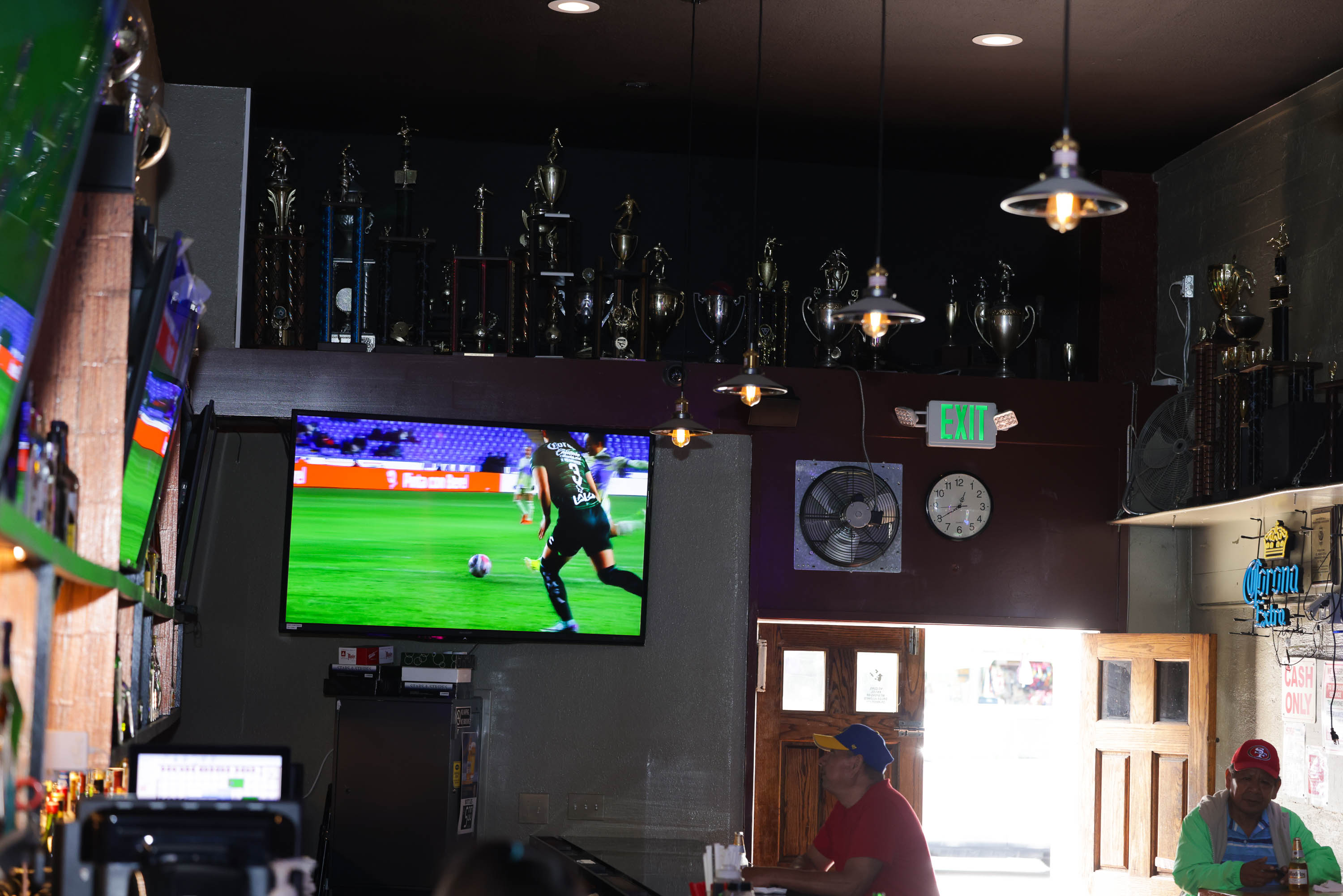 A dimly lit bar with a TV showing soccer, patrons seated, trophies displayed, and an &quot;EXIT&quot; sign illuminated.