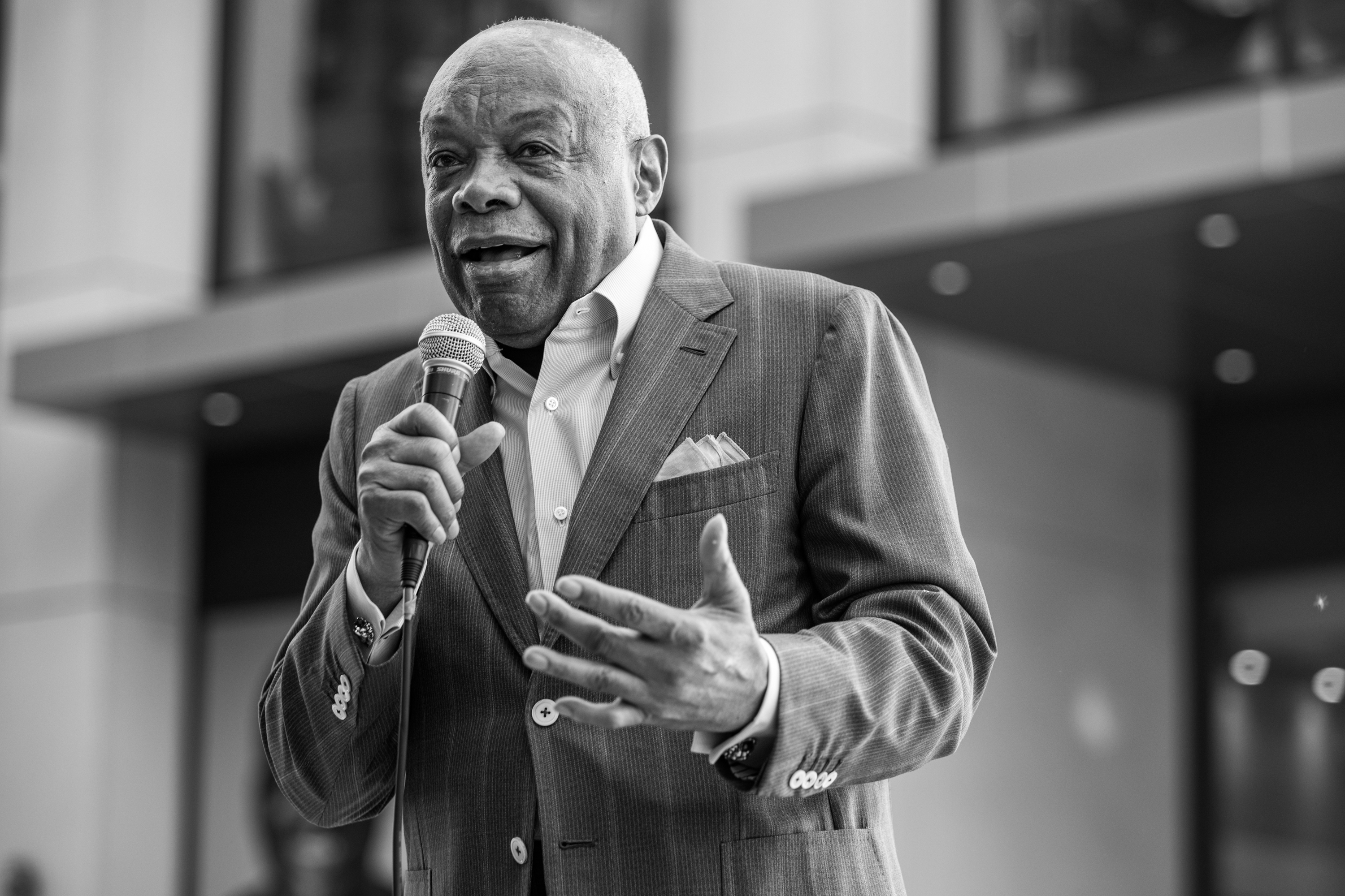 A Black man in a suit speaks into a microphone, gesturing emphatically. The image is black and white.