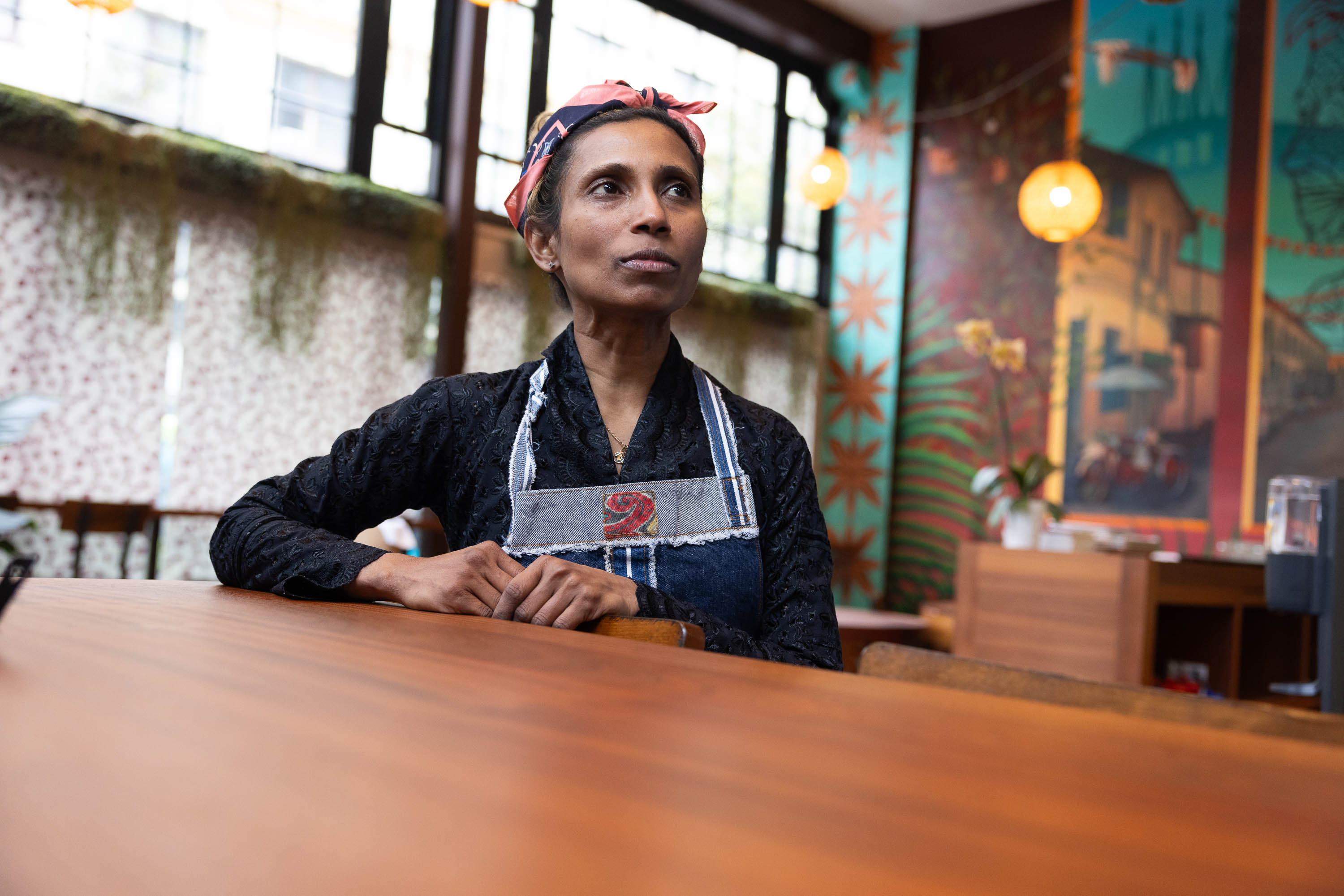 A woman in an apron stands behind a table in a colorful cafe, looking thoughtfully to the side.
