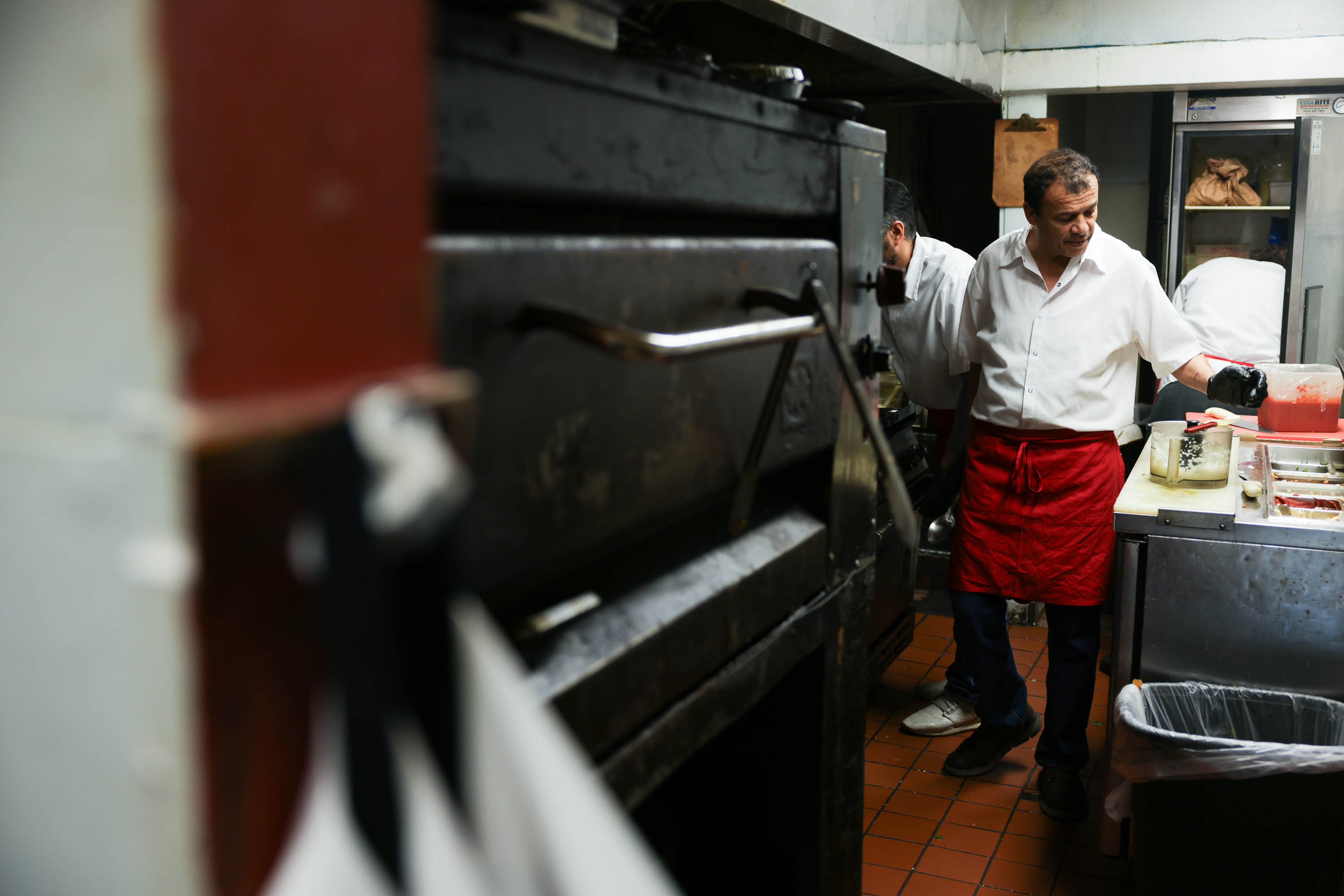 A man in a white shirt and red apron works in a restaurant kitchen near a large oven.