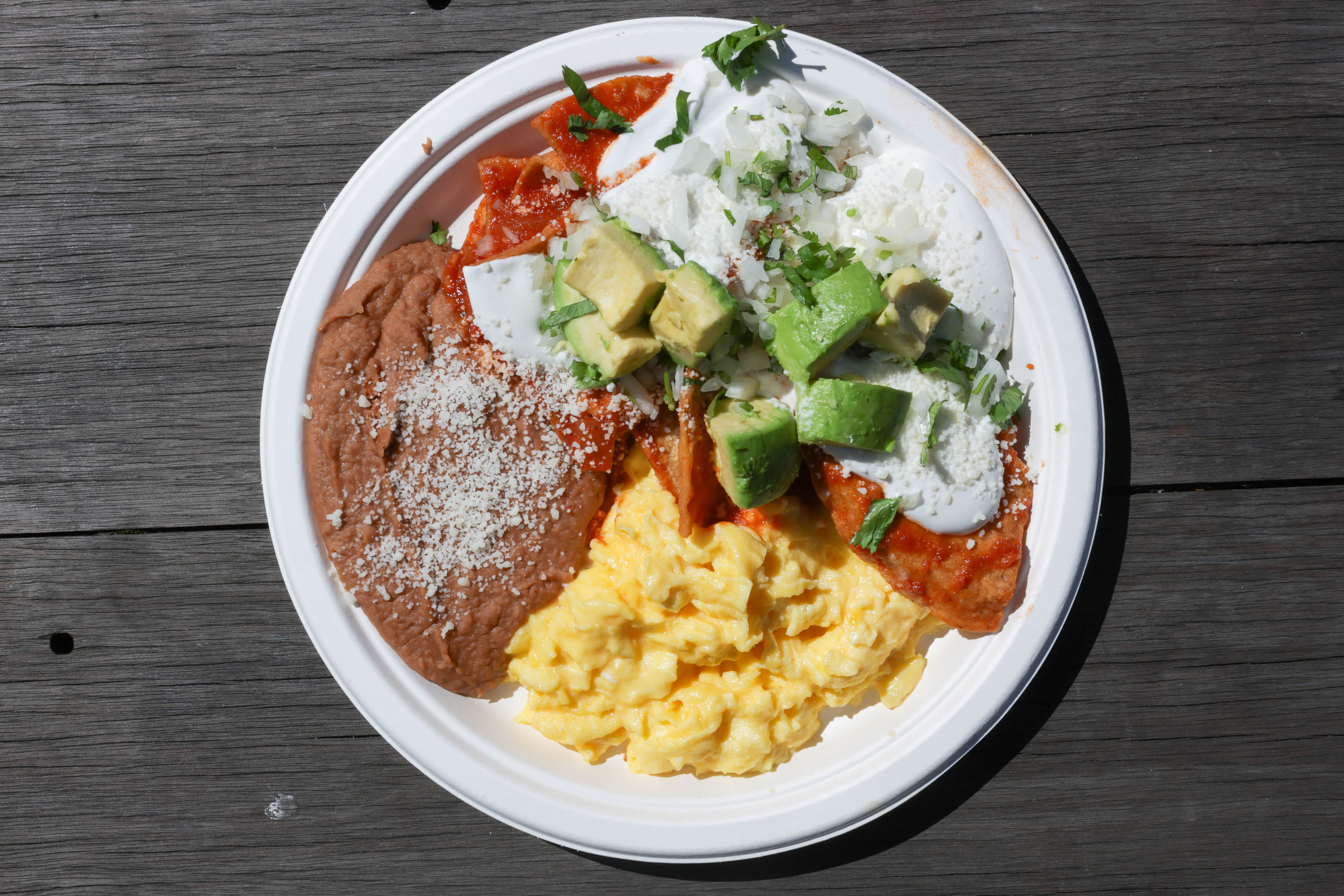 A plate of food with scrambled eggs, refried beans, cheese-topped enchilada, diced avocado, and sour cream.
