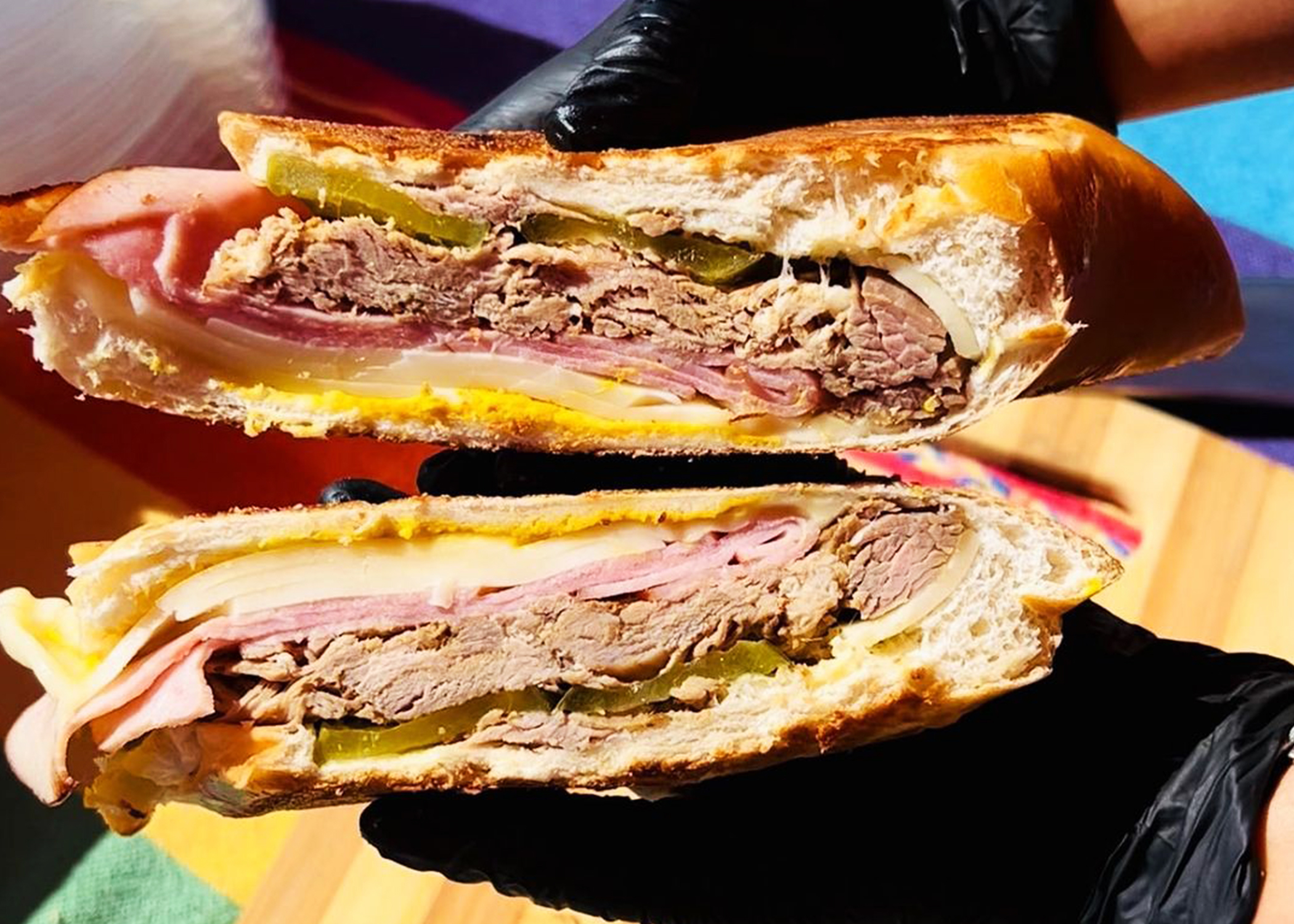 A hand in a black glove holds a meat and cheese sandwich, cut to reveal layers inside.