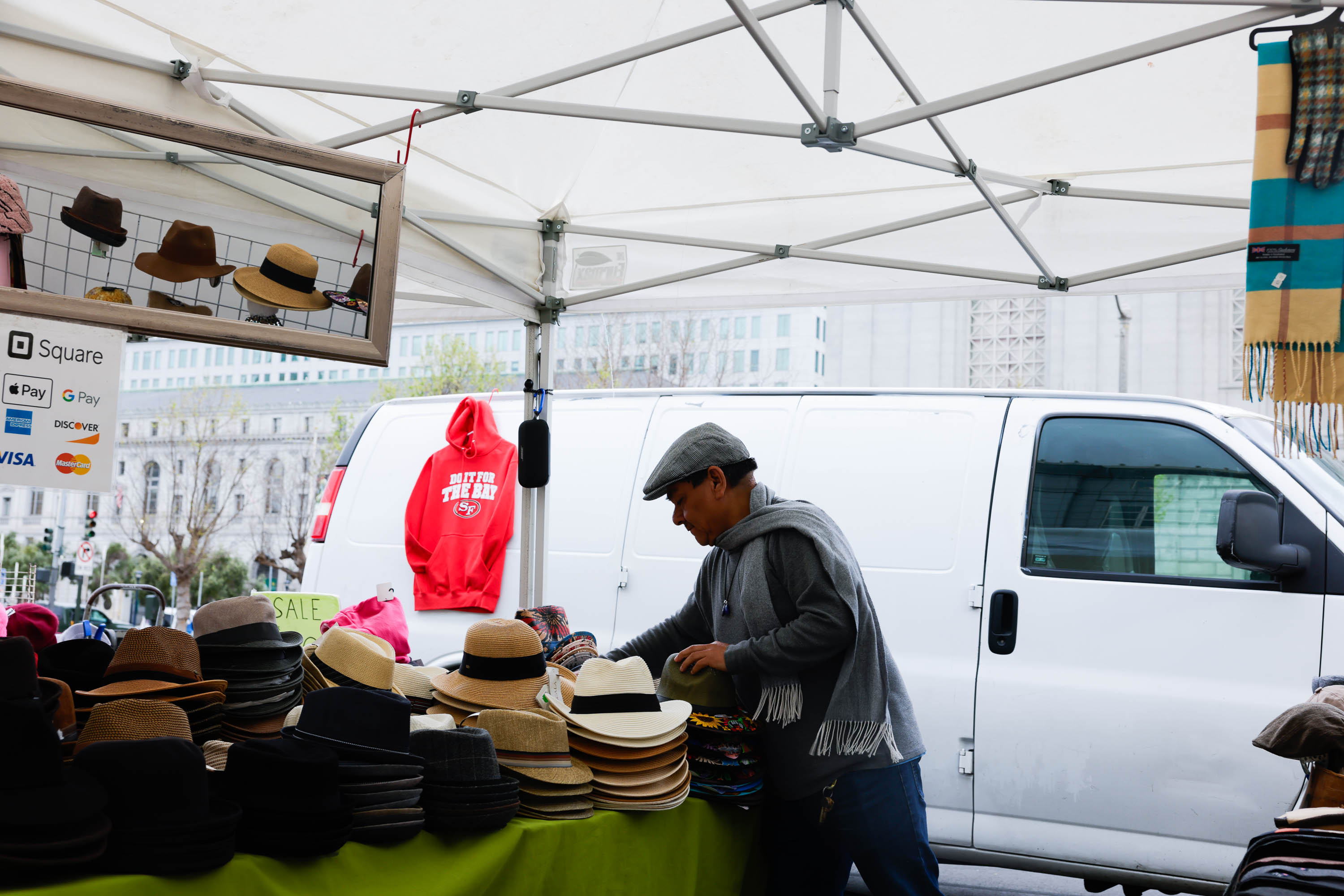a Latino man under a canopy sets up a merchandise table in front of a white van