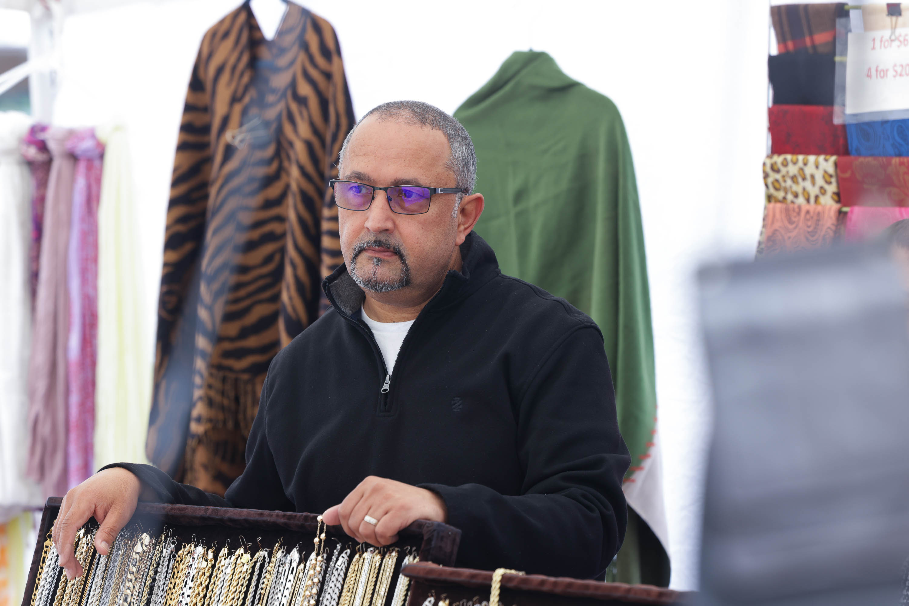 a middle-aged middle eastern man in glasses frowns at a market stall surrounded by apparel