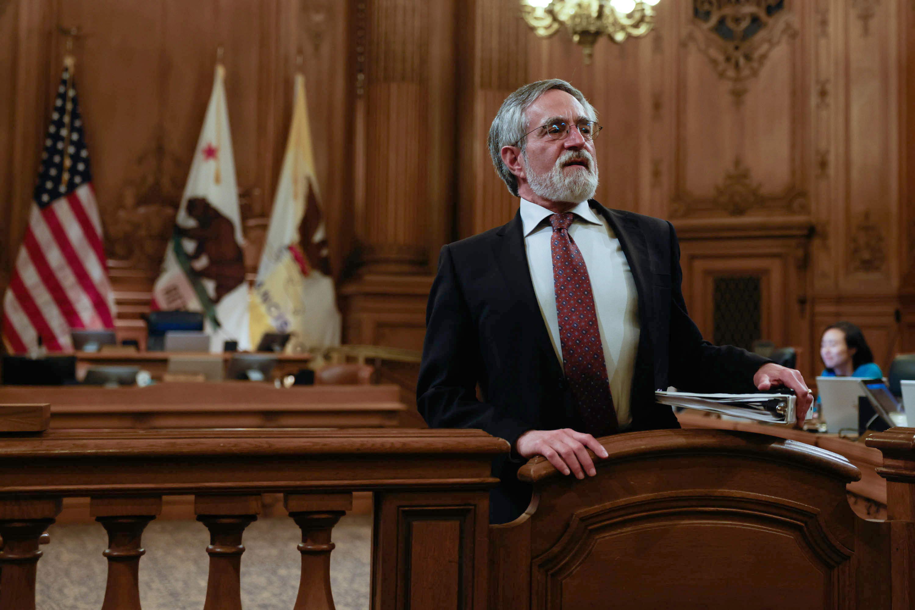A man stands at a podium in a grand room with flags and a person working in the background.