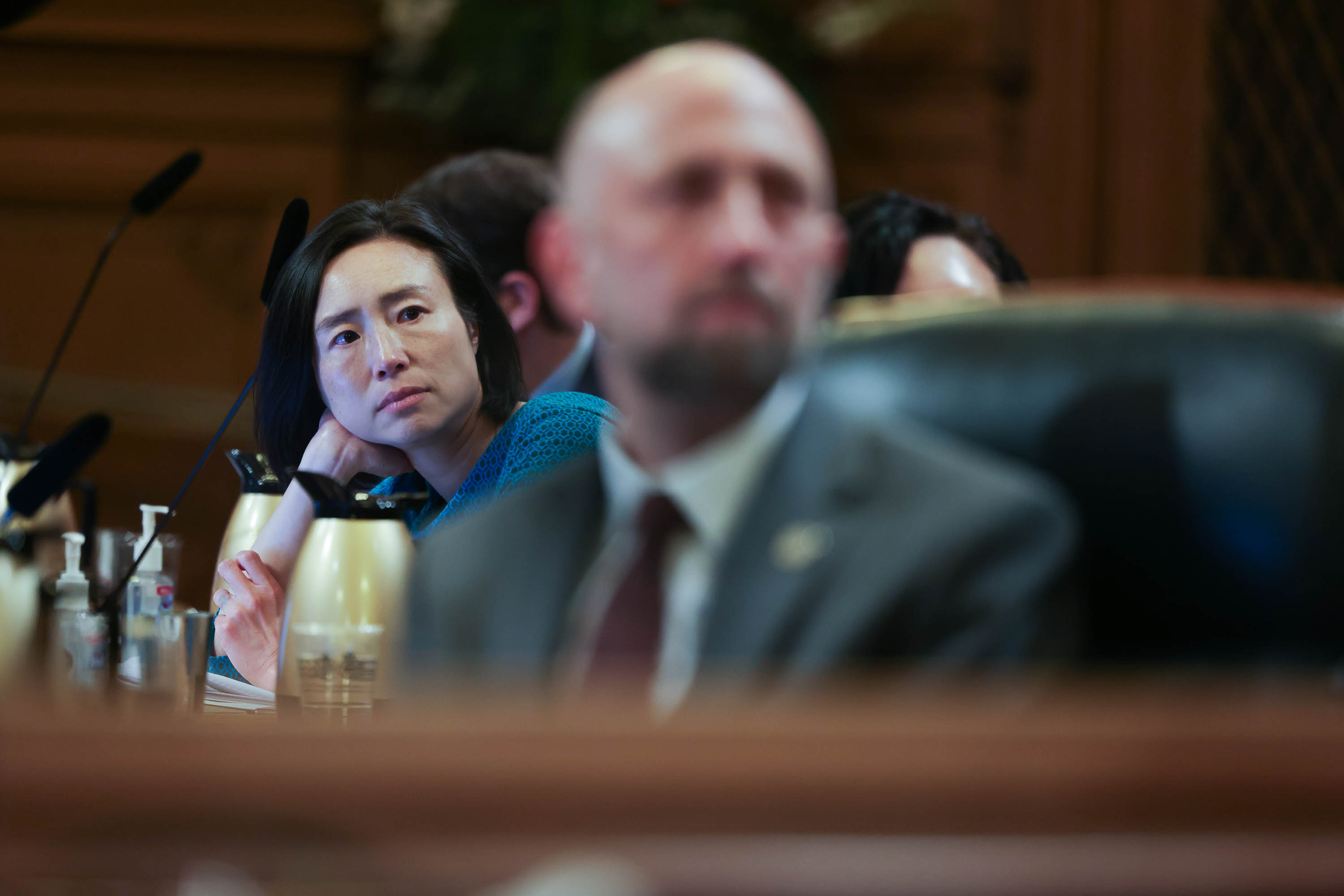 A woman in a hearing room looks pensive, surrounded by microphones and blurred foreground objects.