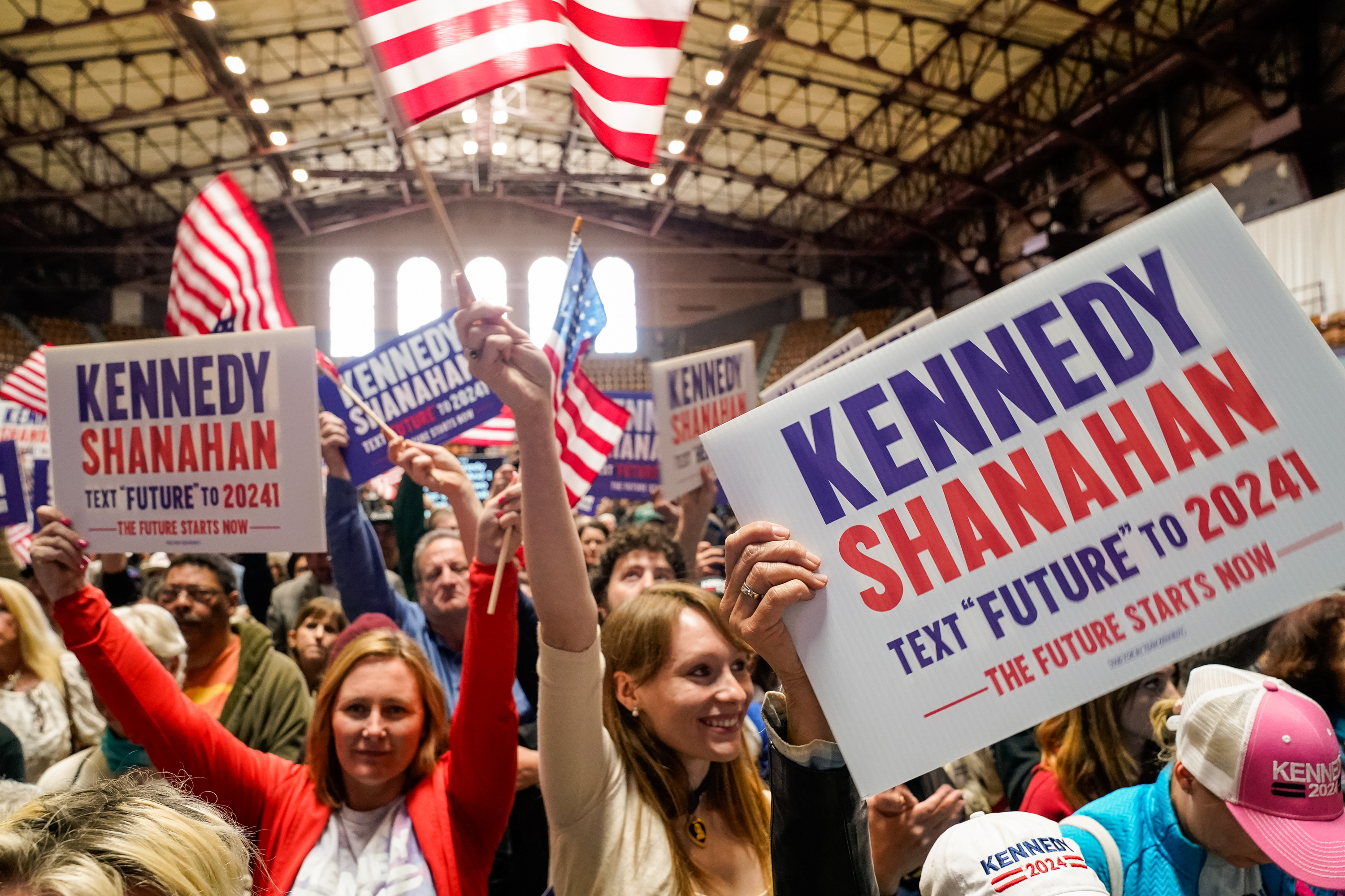 A crowd with &quot;KENNEDY SHANAHAN&quot; political signs, American flags, and a gymnasium backdrop.