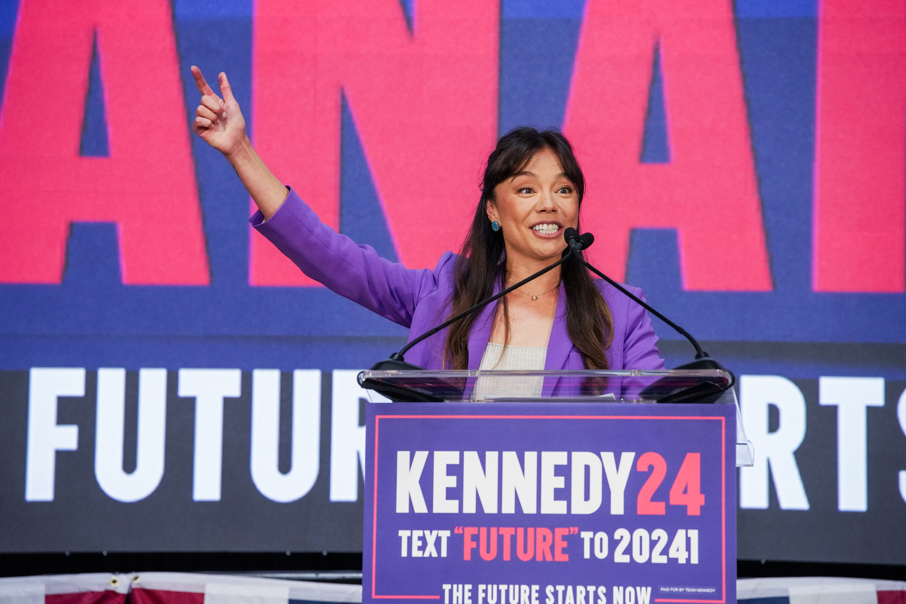 A smiling person in a purple suit behind a podium with &quot;KENNEDY 2024,&quot; raises a peace sign, with a &quot;FUTURE&quot; backdrop.