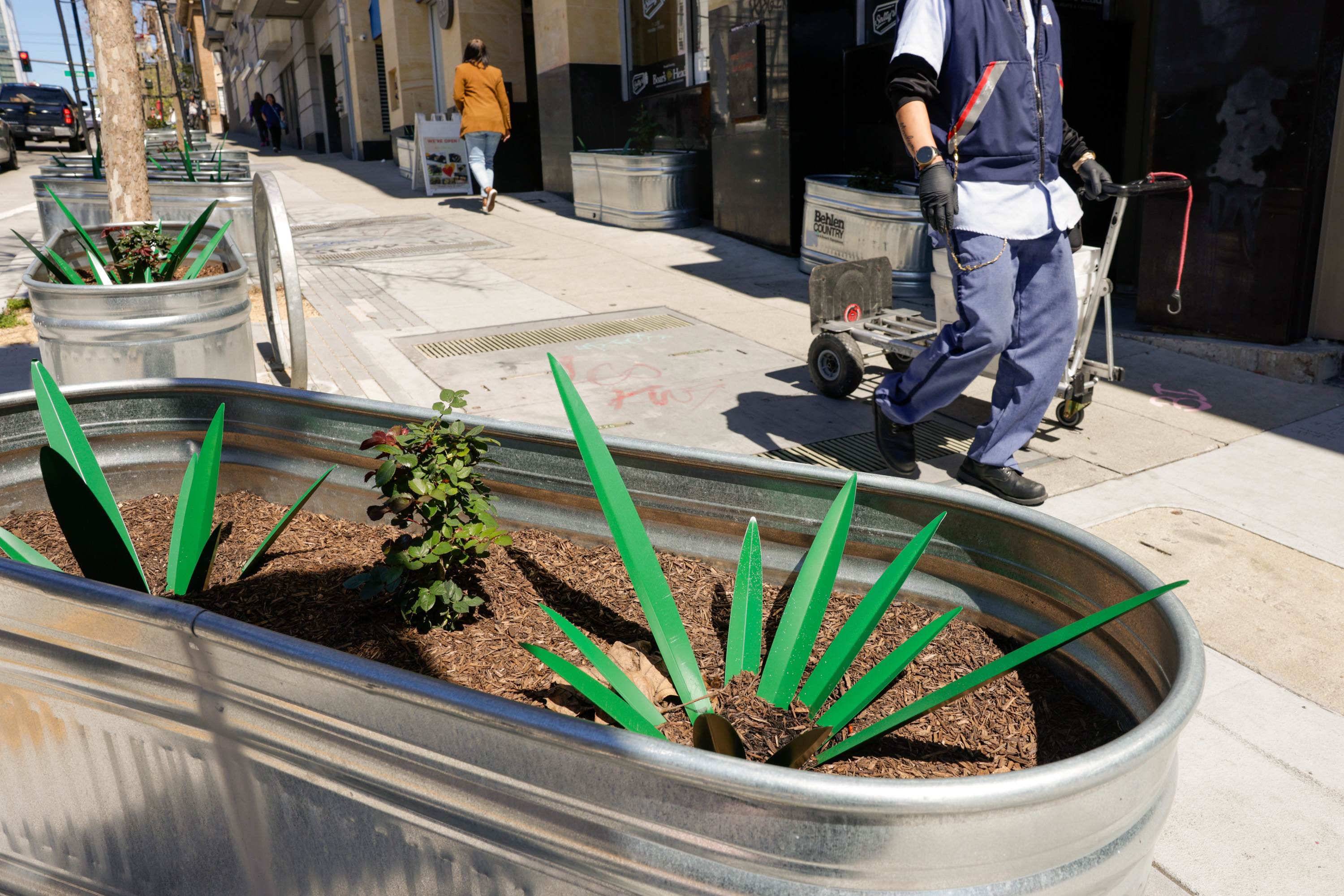 Metal planters with greenery line a sunny sidewalk; a person in work attire walks by, pulling a hand truck.
