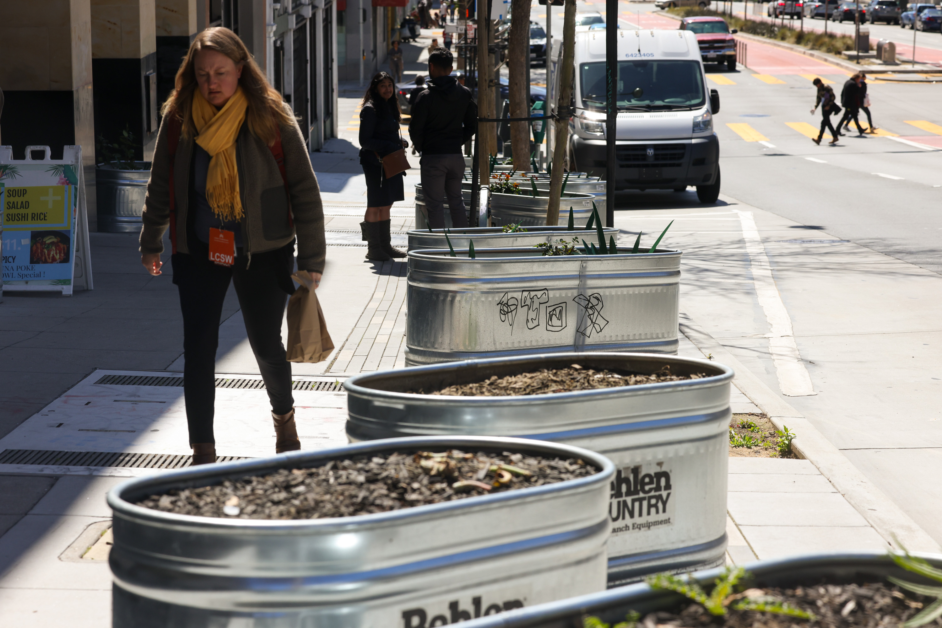 A sidewalk with people walking past large metal planters under a clear sky.