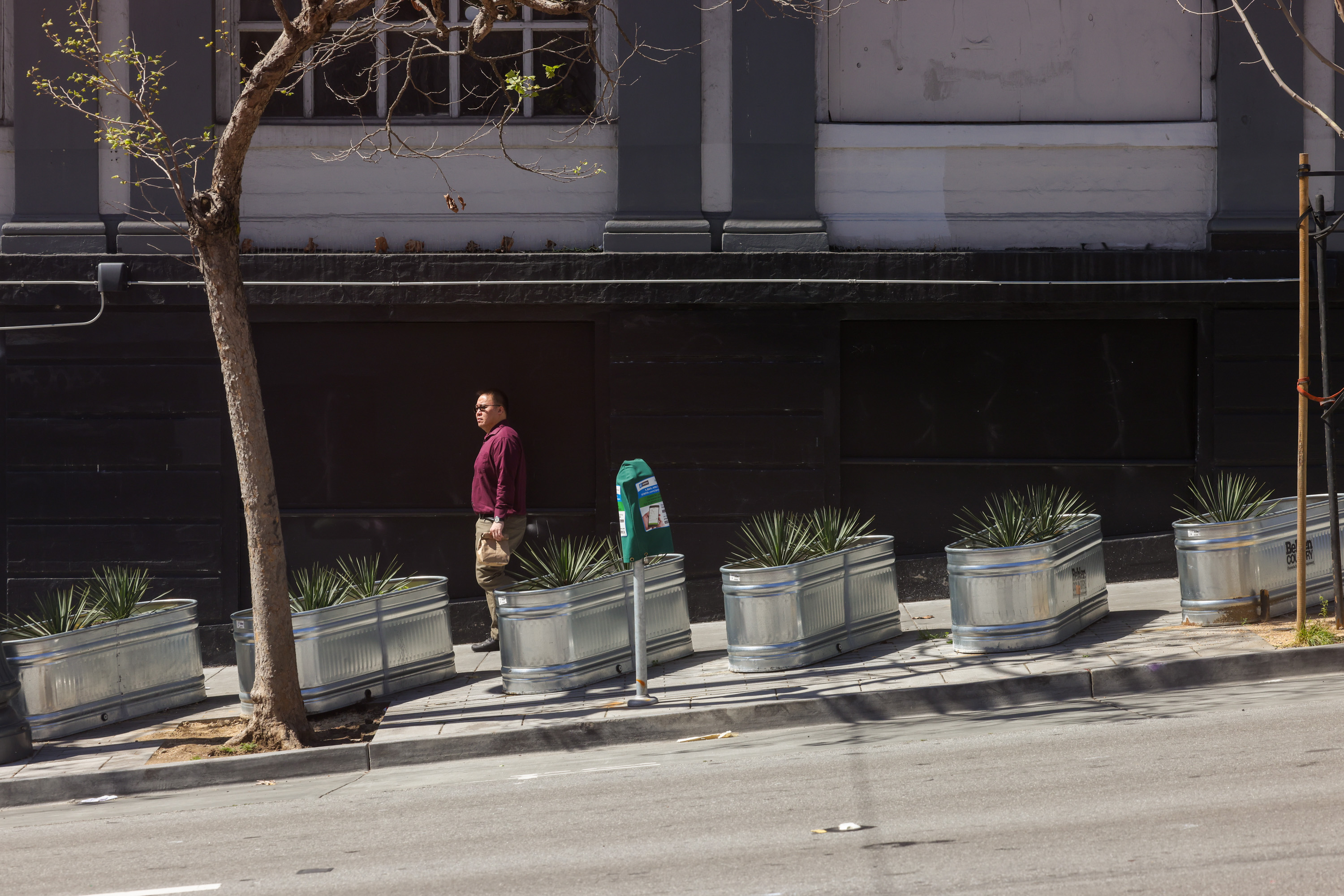 A person walks by metal planters on a sunlit urban sidewalk.