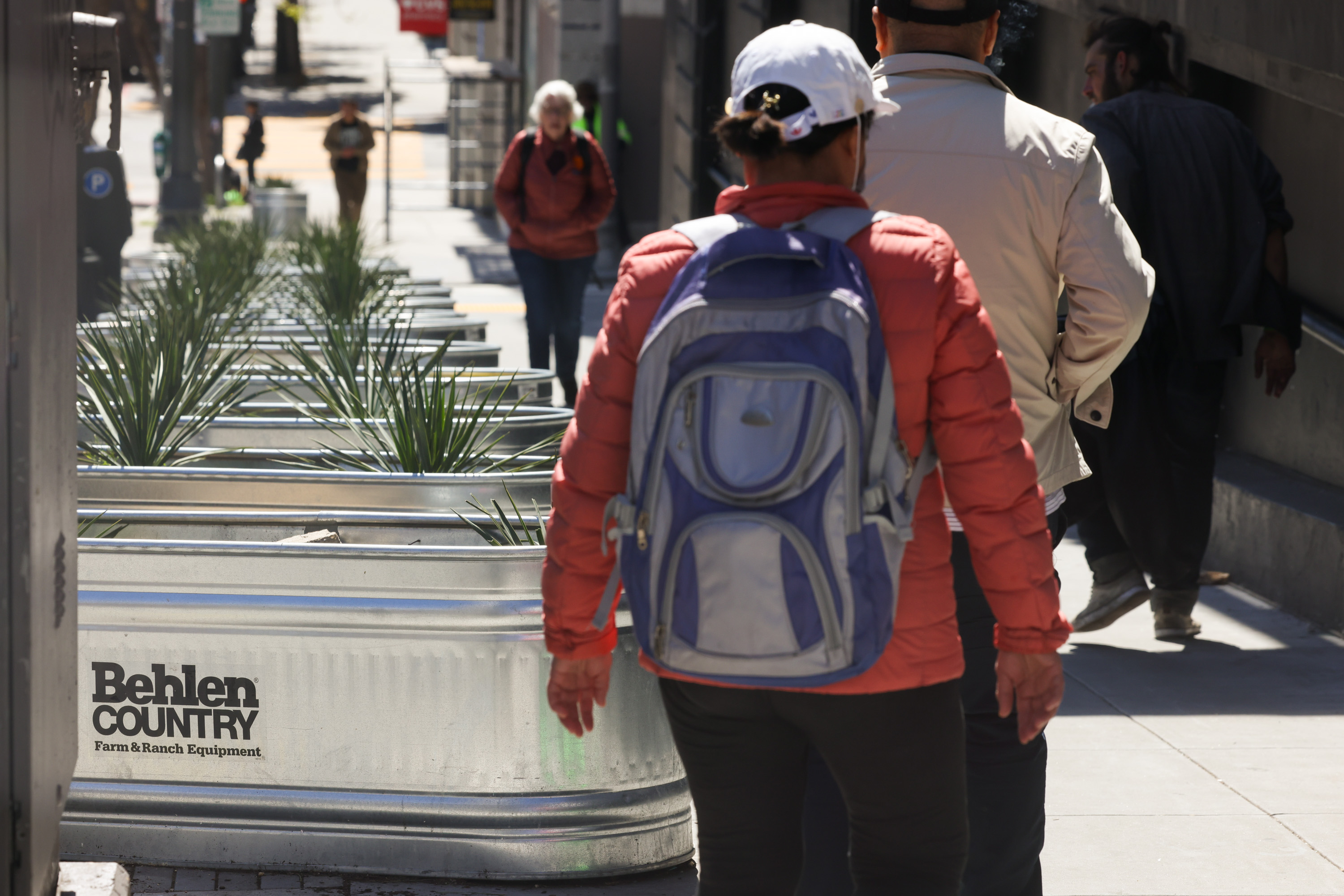 A person with a backpack walks by metallic planters on a sunny, urban sidewalk. Other pedestrians are visible in the background.