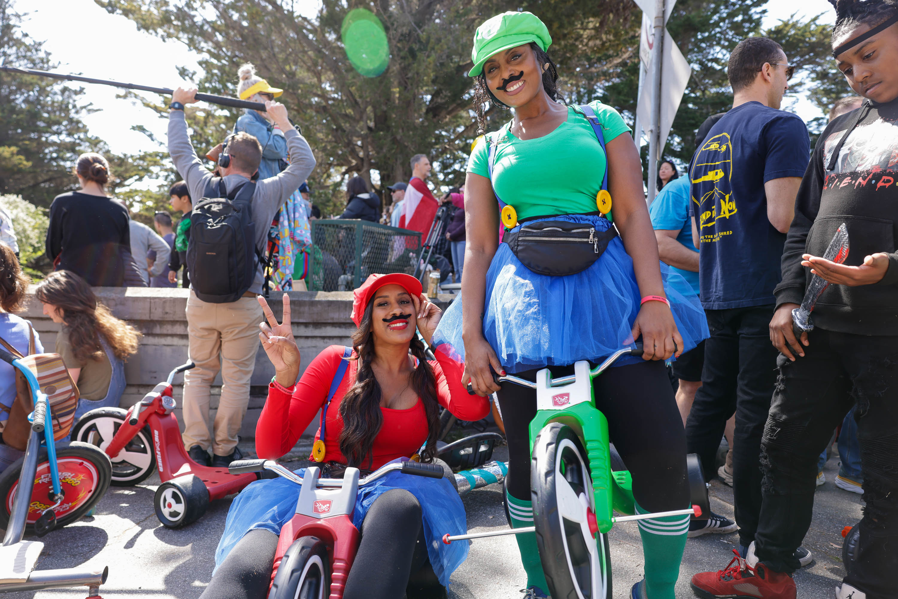 Two people dressed as Mario characters are riding tricycles; the scene appears to be outdoors at an event.