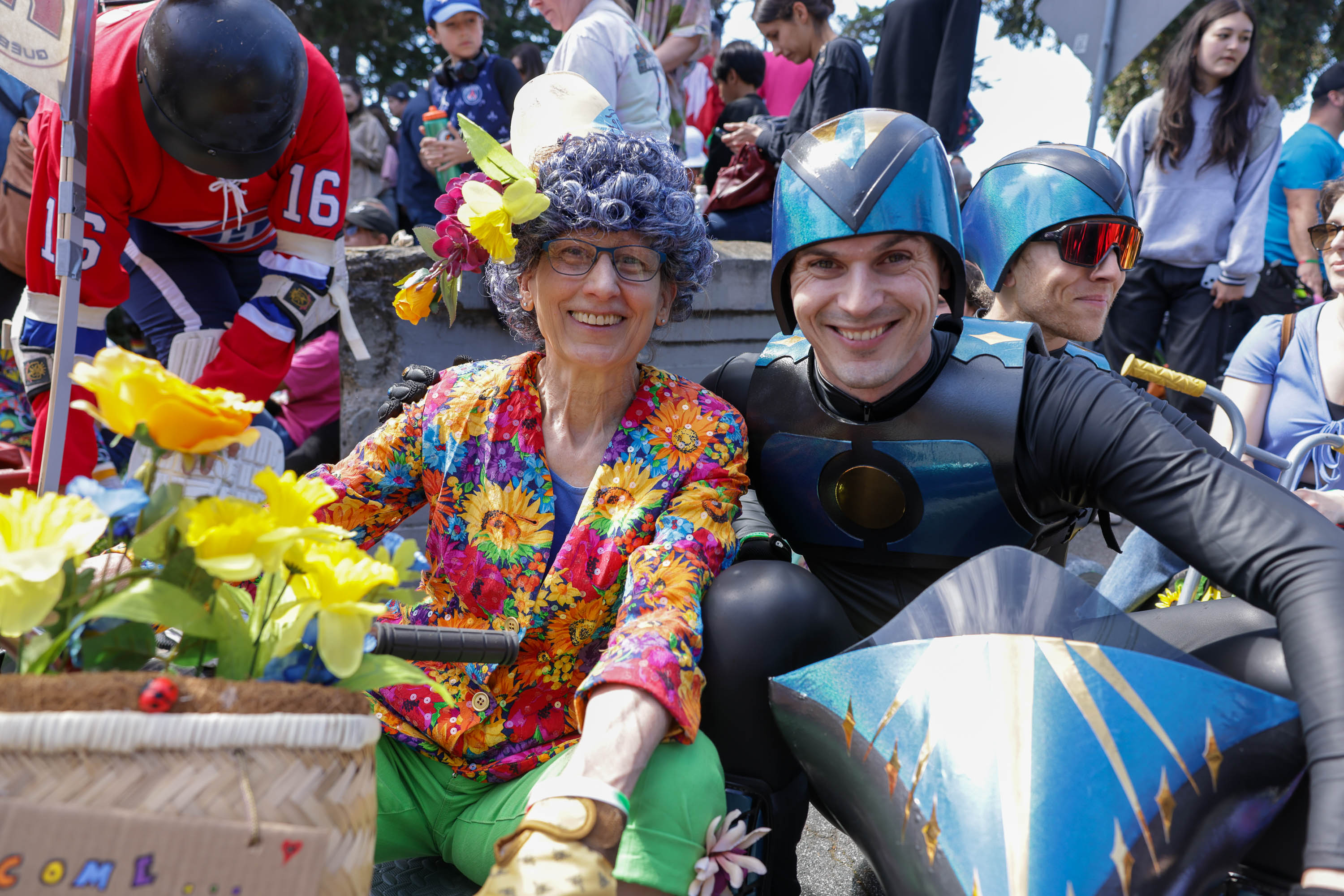 Two people in colorful costumes pose on a flower-adorned bicycle; one is dressed as a superhero, the other in a vibrant floral outfit.