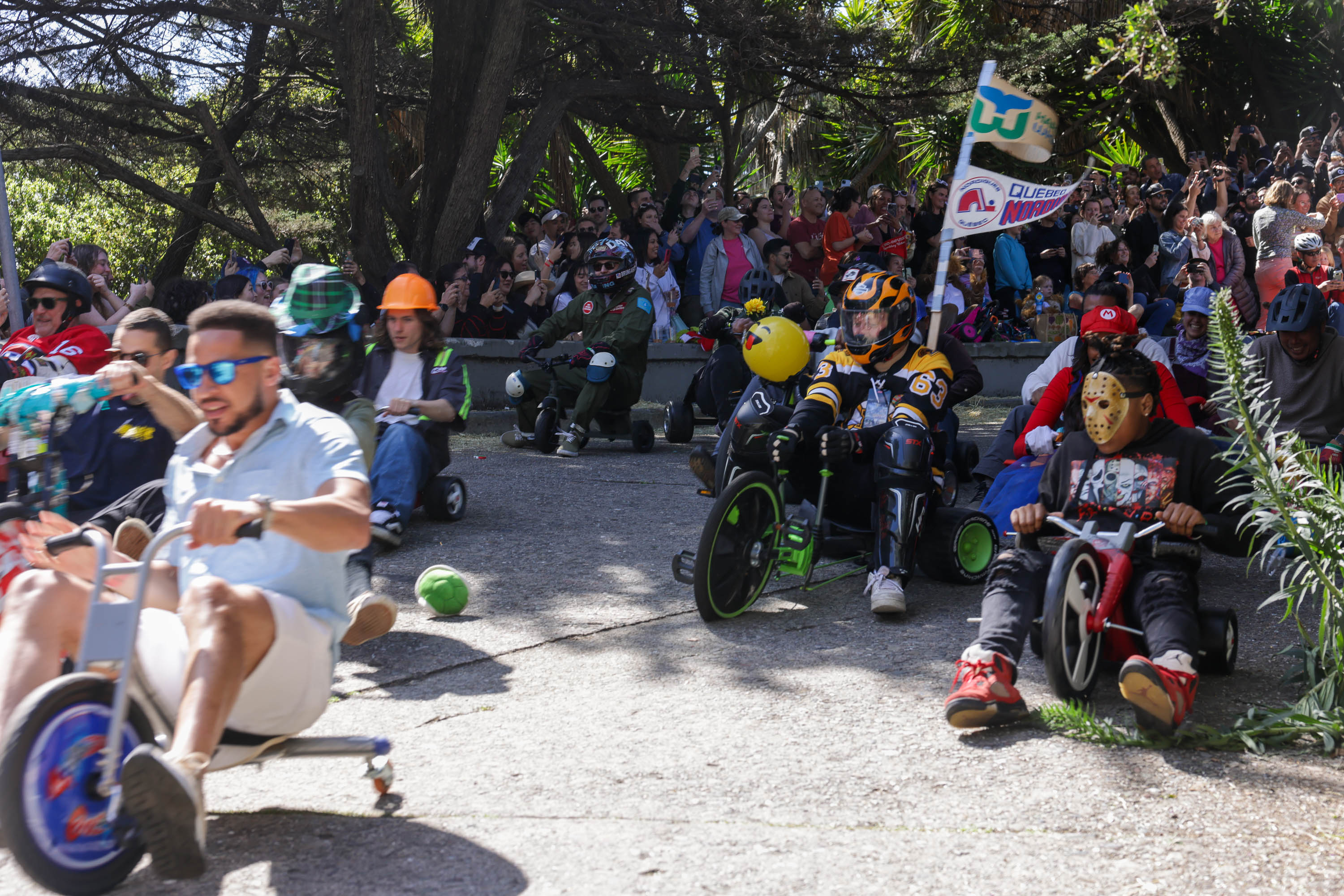 Participants in homemade carts race downhill as a crowd watches. Some wear costumes and helmets, stirring excitement.