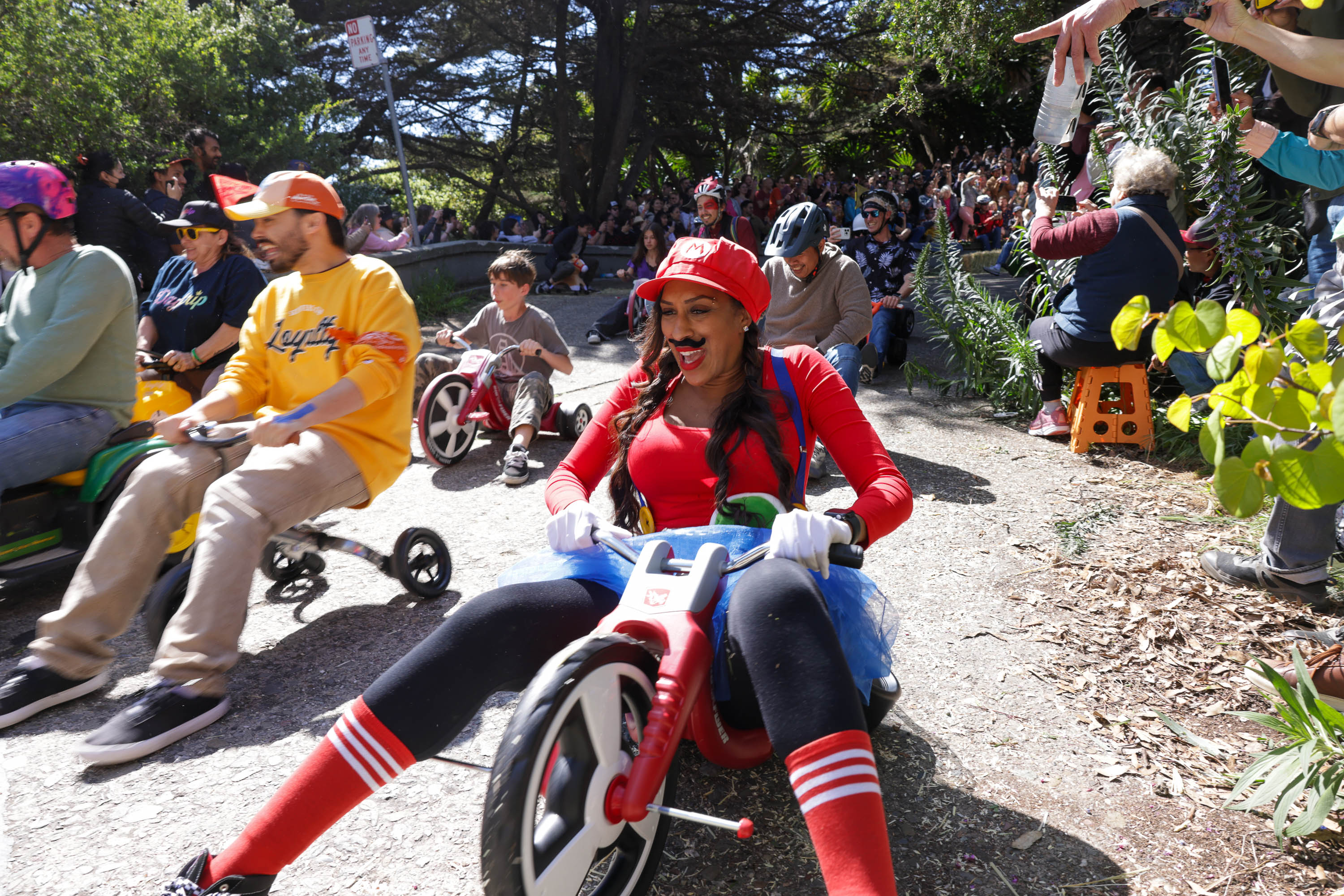 Adults in costumes ride big wheel bikes downhill, with a lively crowd watching.
