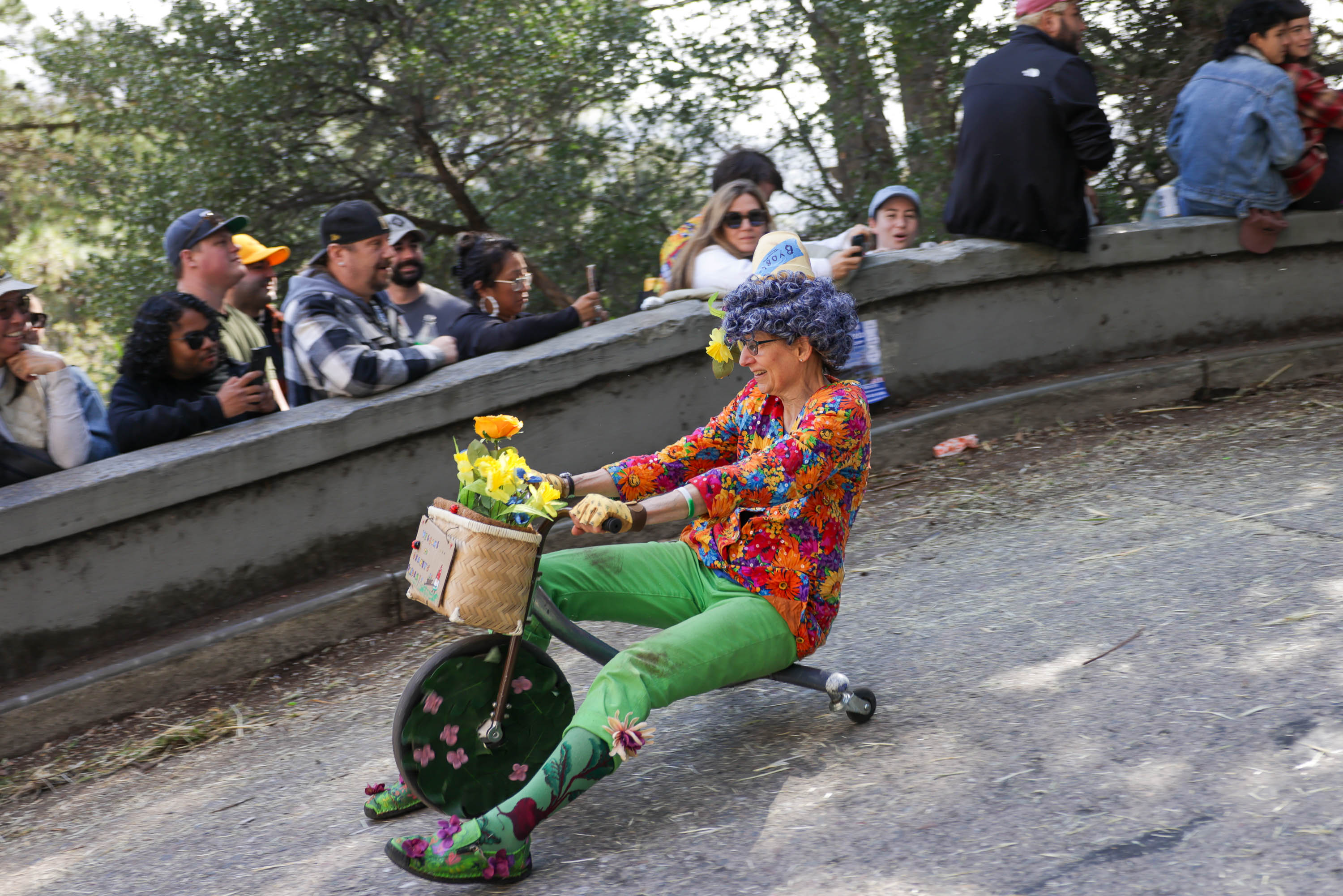A person rides down a hill on a small tricycle, wearing bright attire with flowers, as onlookers in the background watch.