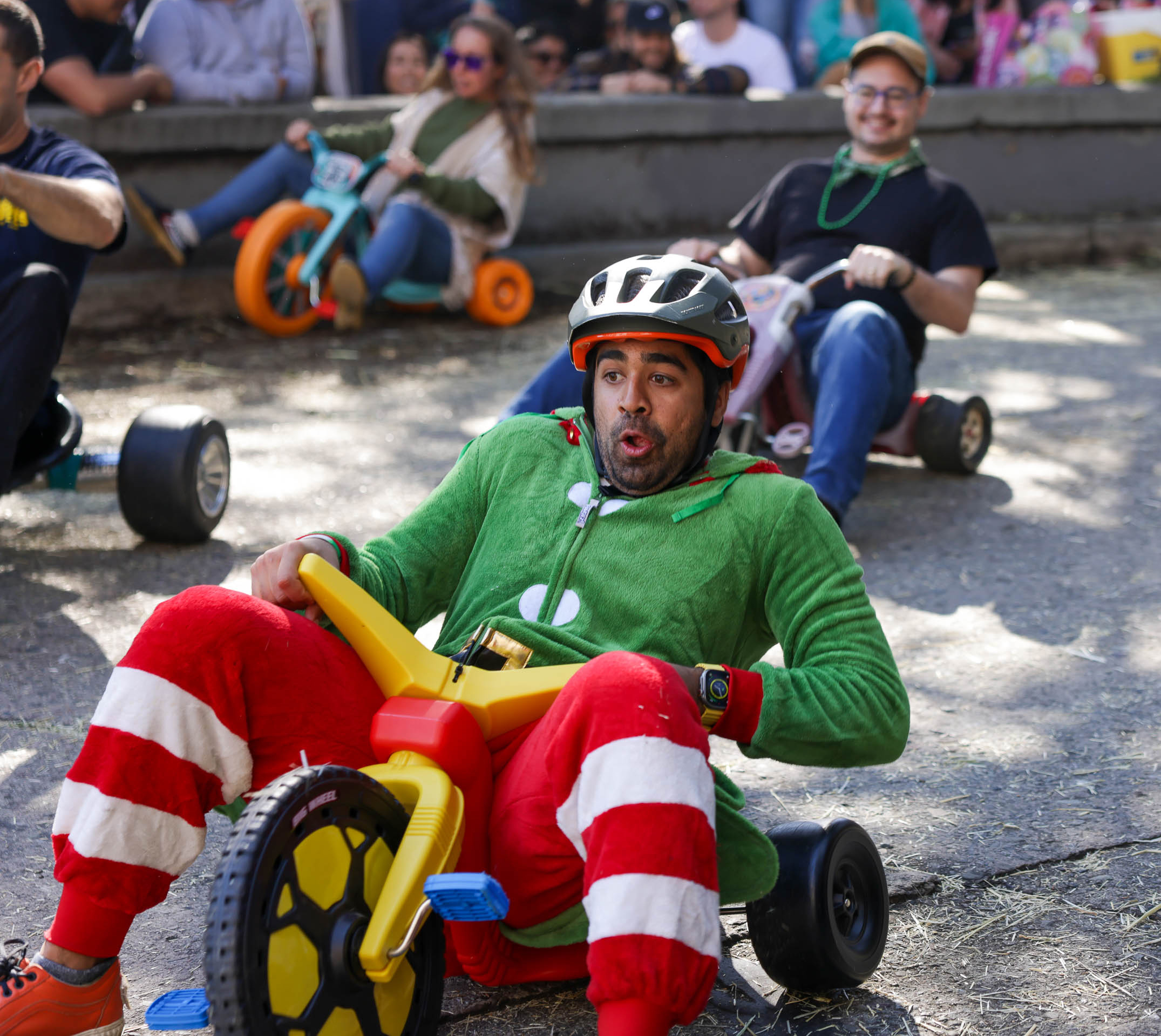 A person in an elf costume races downhill on a toy tricycle, looking amused, as onlookers watch.
